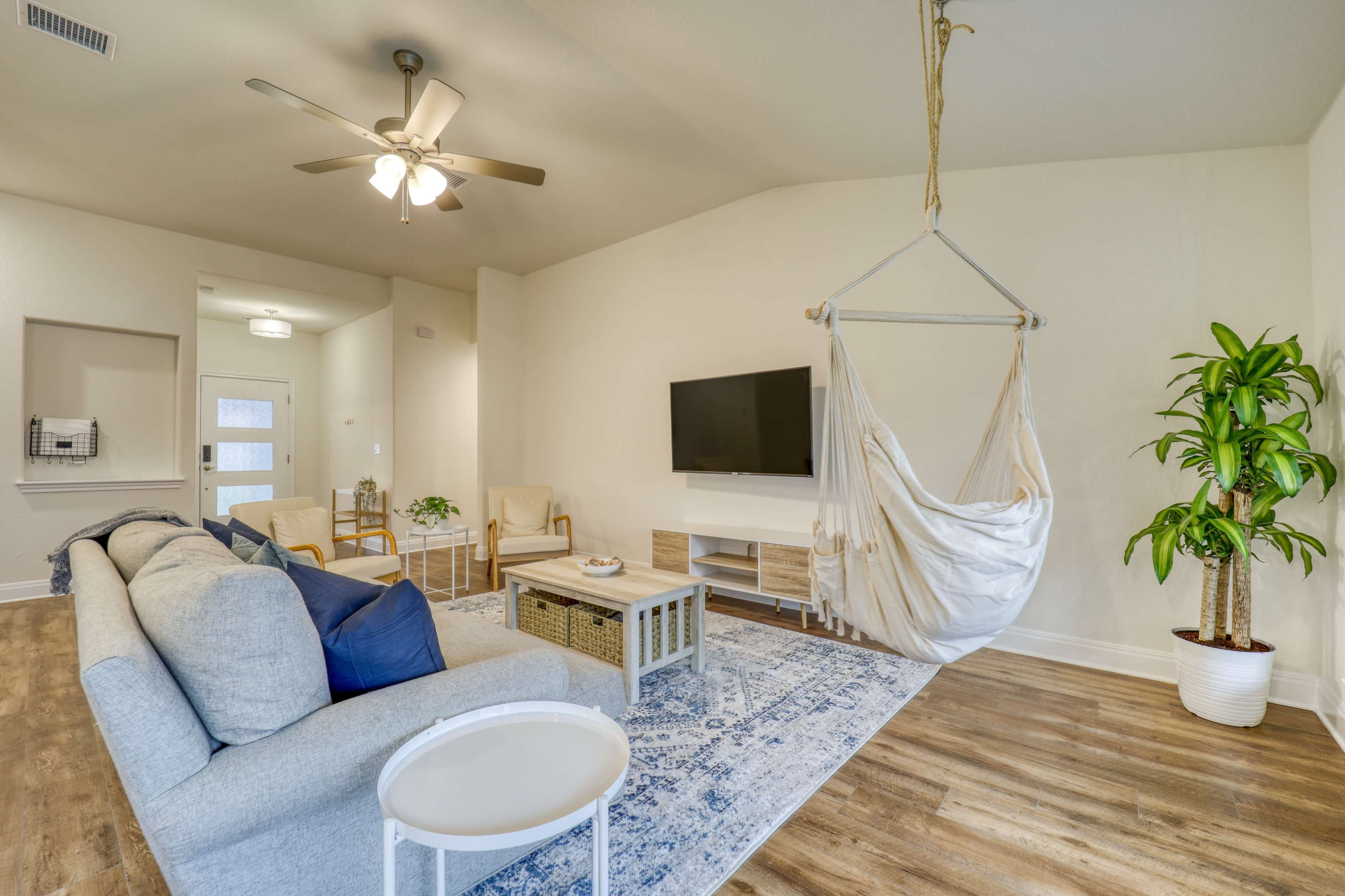 A modern living room with a light-colored sofa, a television mounted on the wall, a hanging chair, and a potted plant.