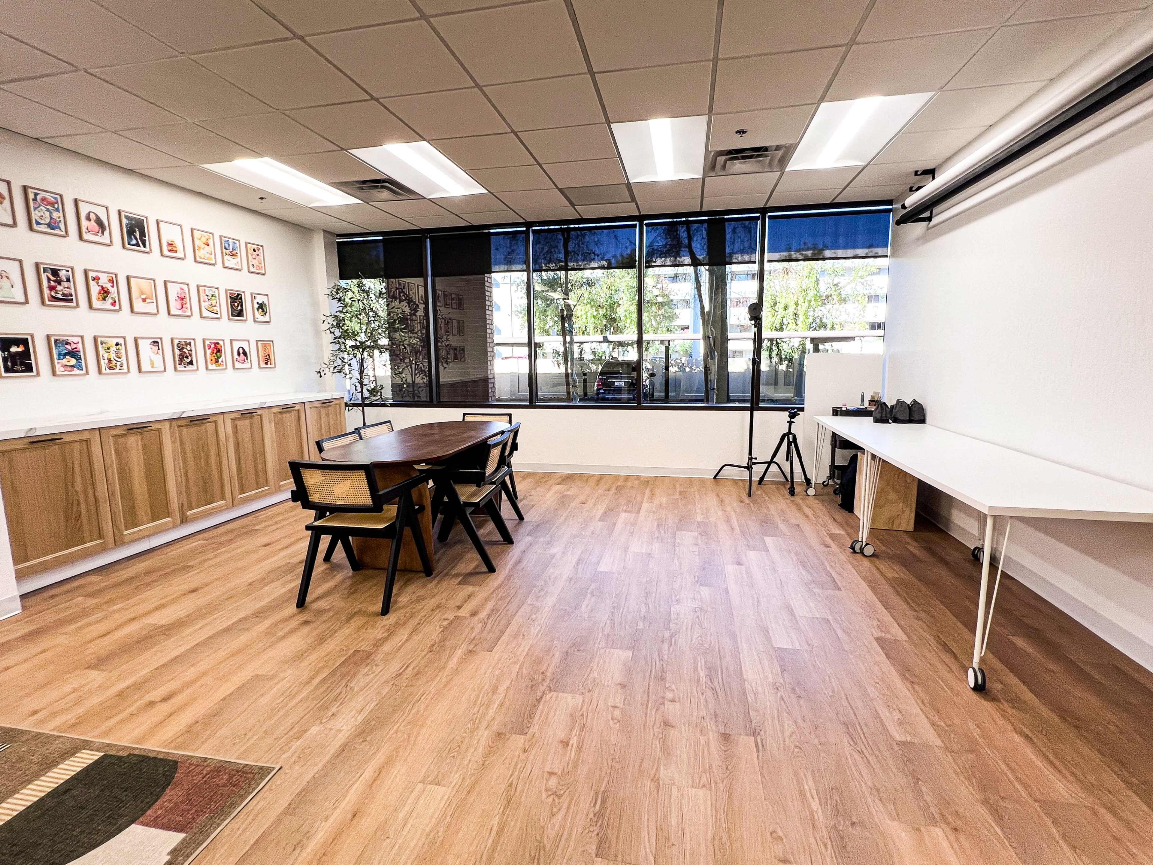 A bright, spacious room with a wooden table, chairs, and large windows displaying an outdoor view.