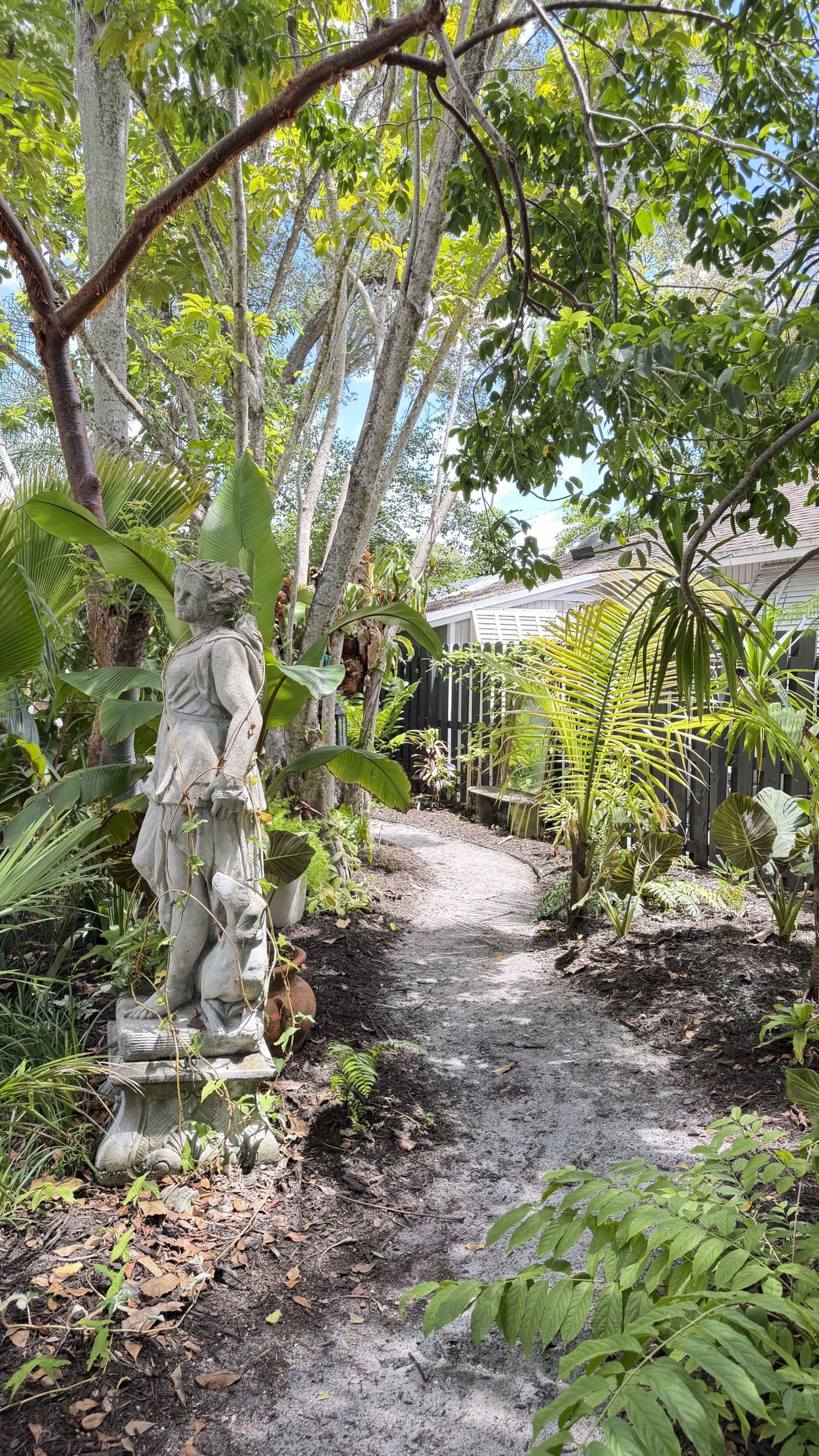 A stone statue stands next to a winding path surrounded by lush greenery and tropical plants.