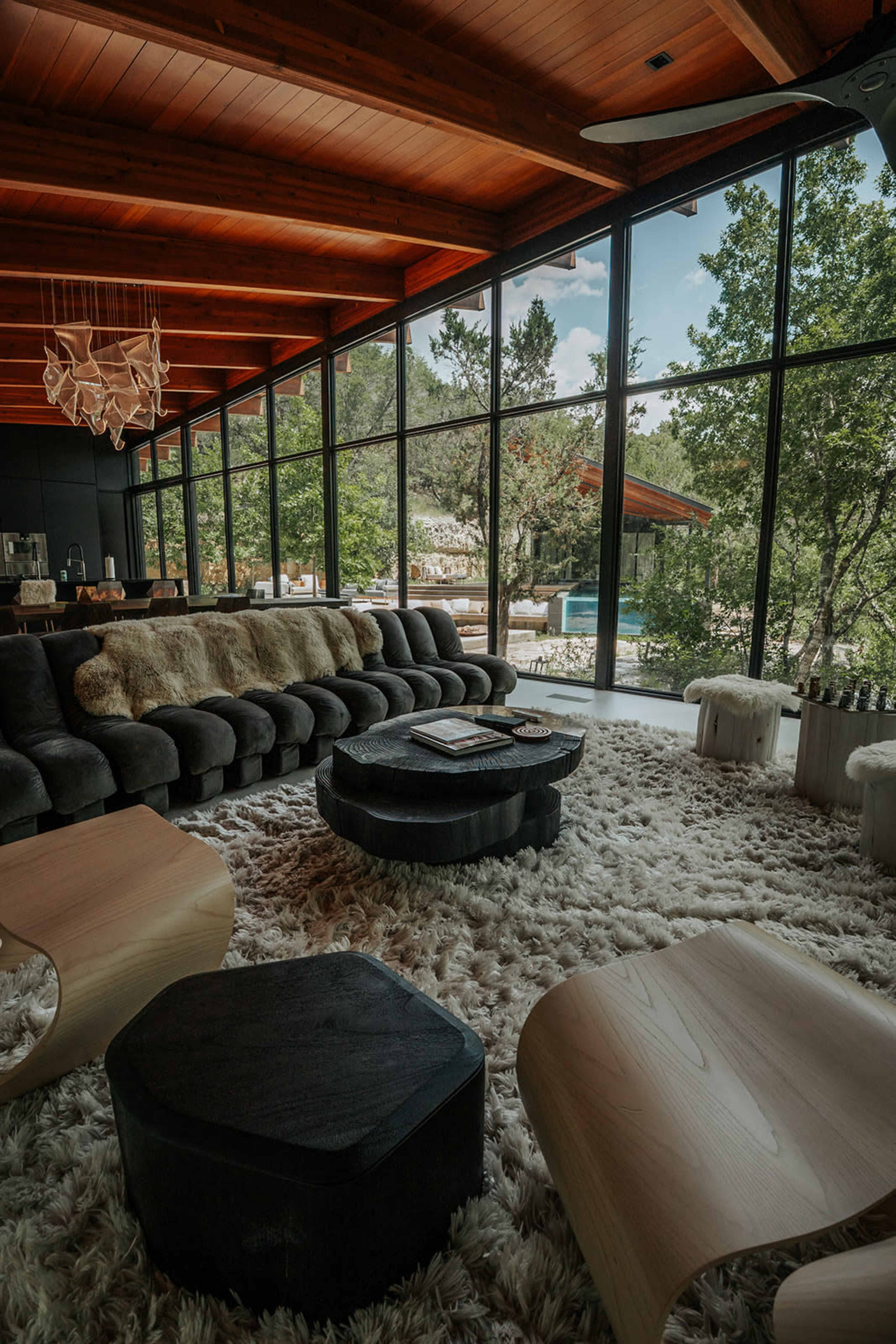 The image shows a modern living room with large windows overlooking a natural landscape, featuring a plush sectional sofa, unique coffee tables, and a textured rug.