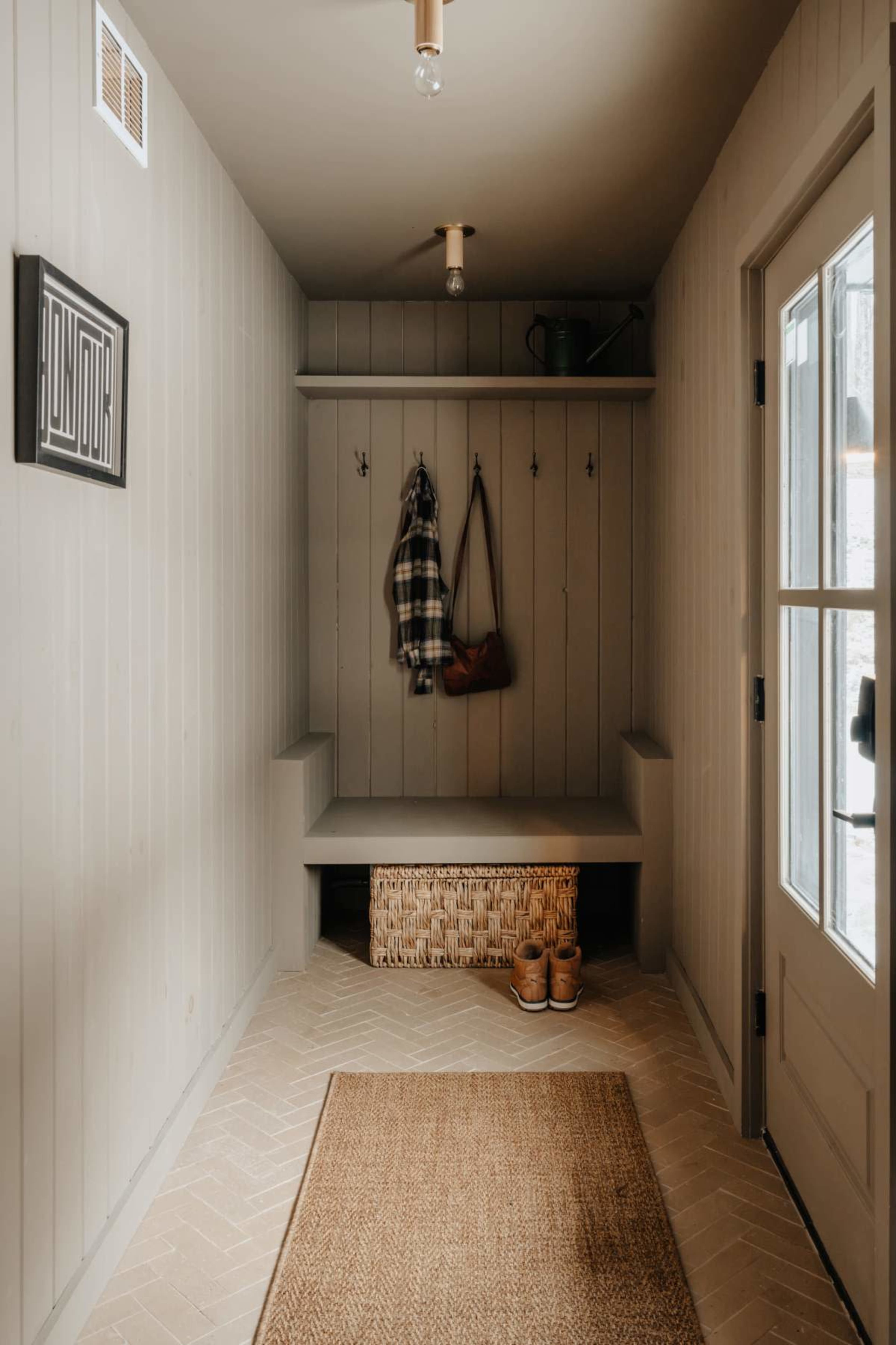 The image shows a minimalist entryway with a bench, wall hooks for hanging coats, a woven storage basket, and a door leading outside.