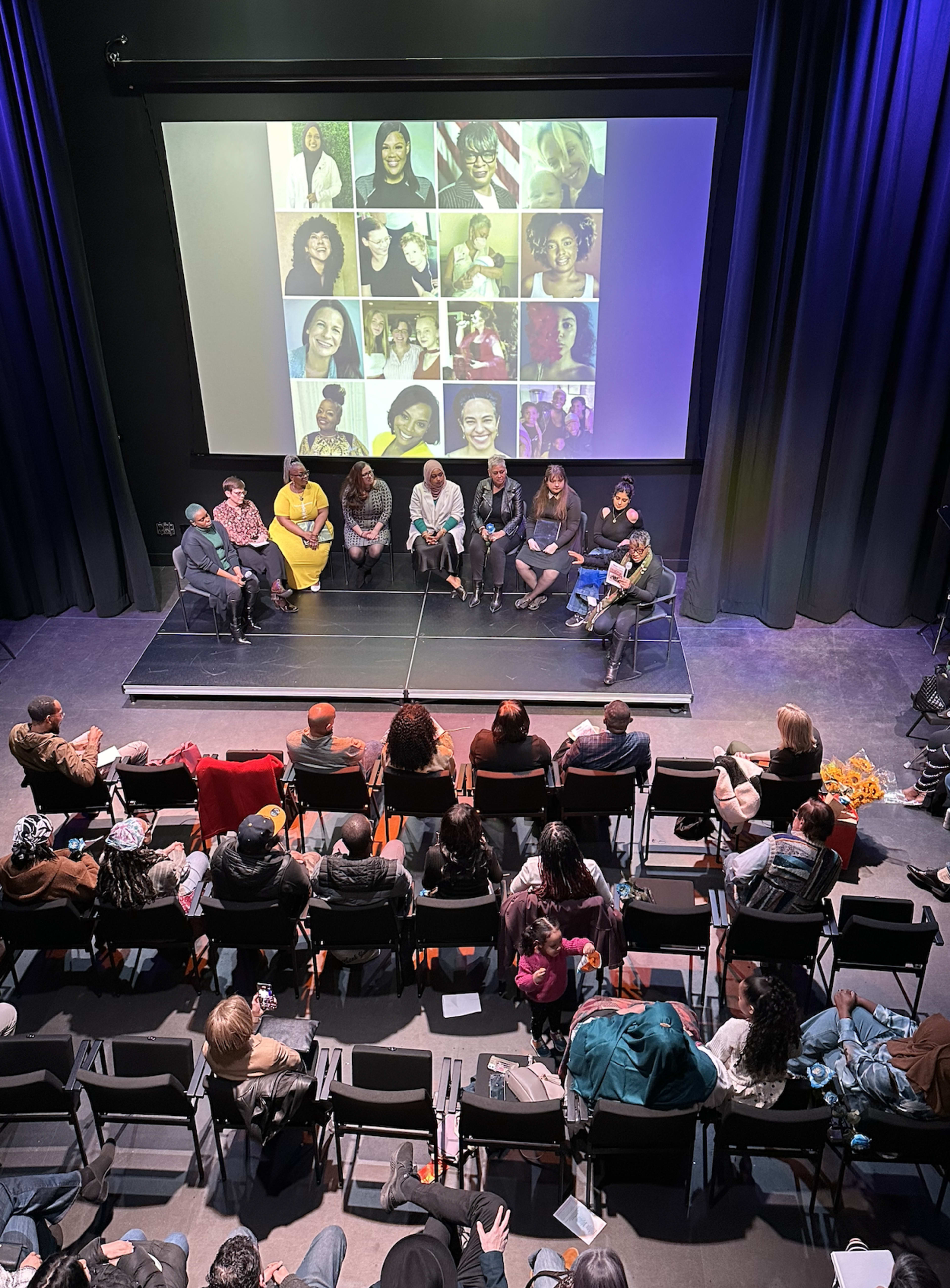 A panel of speakers sits on a stage with a large screen displaying photos behind them, while an audience watches from seated rows.