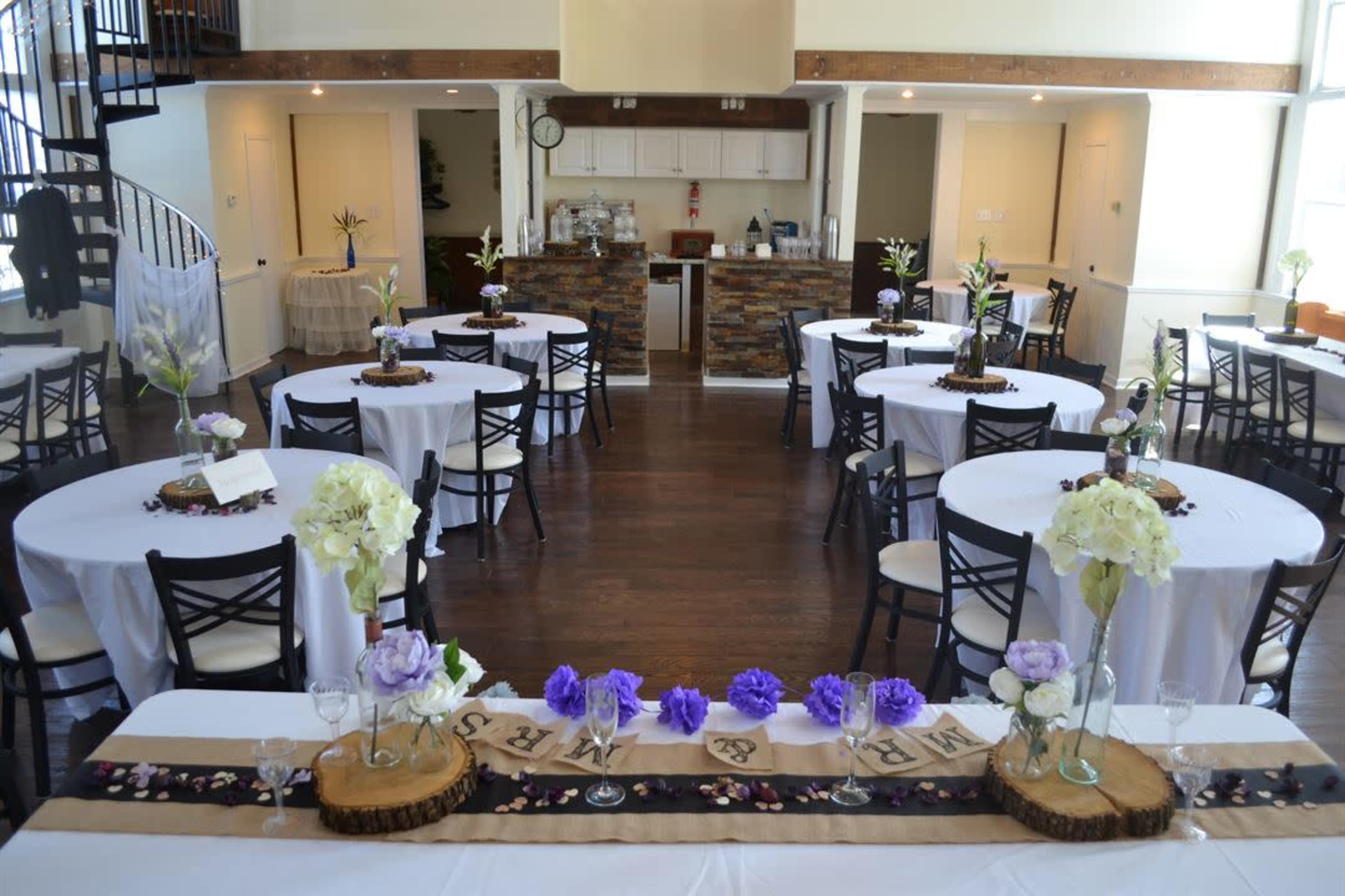 The image shows a decorated event space with circular tables arranged around a central bar area, featuring floral centerpieces and a wooden decorative runner.