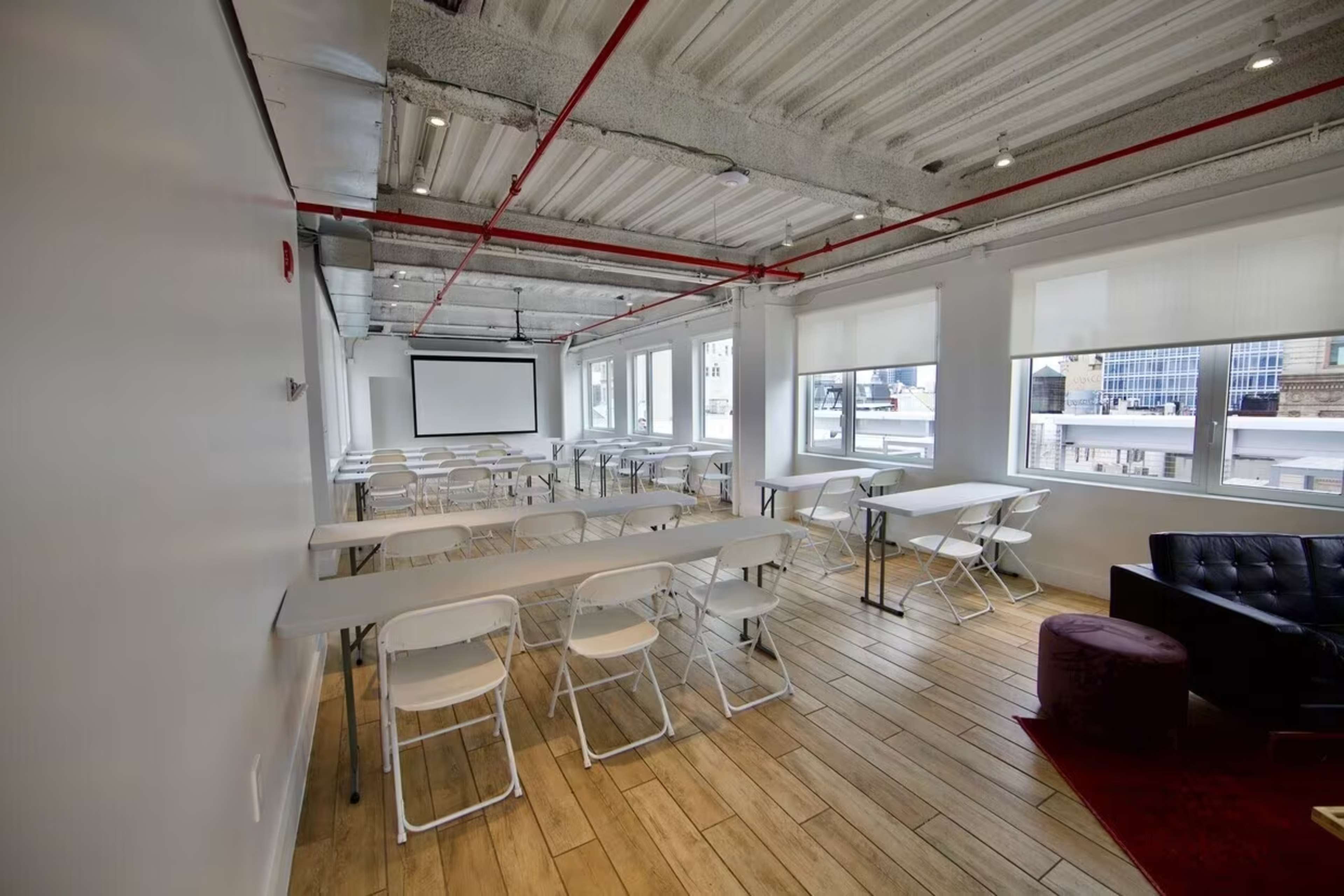 A modern meeting room features several rows of tables with chairs, large windows, and a projector screen.