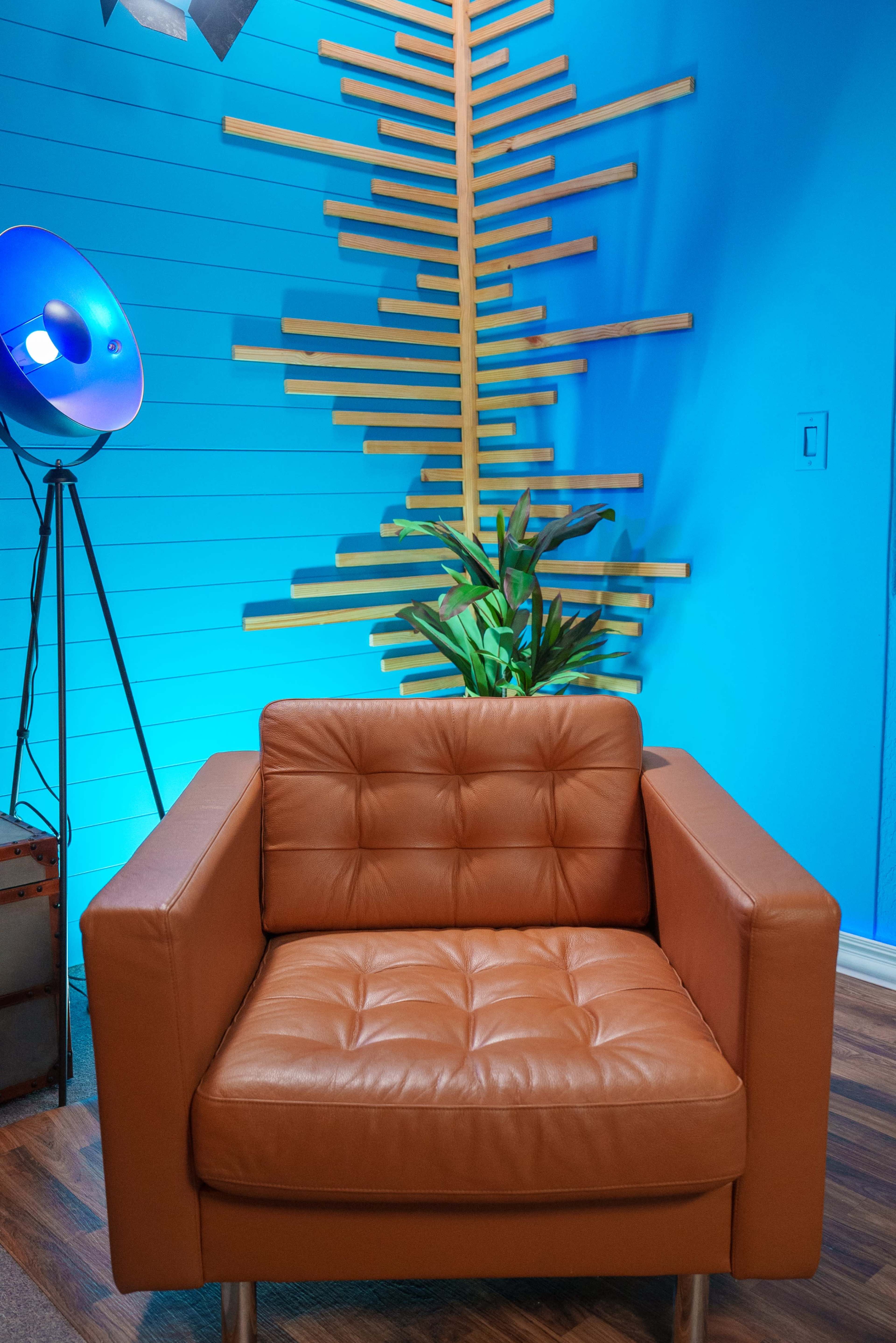 A modern leather armchair in a warm orange hue is positioned beside a tall plant against a bright blue wall decorated with abstract wooden elements.