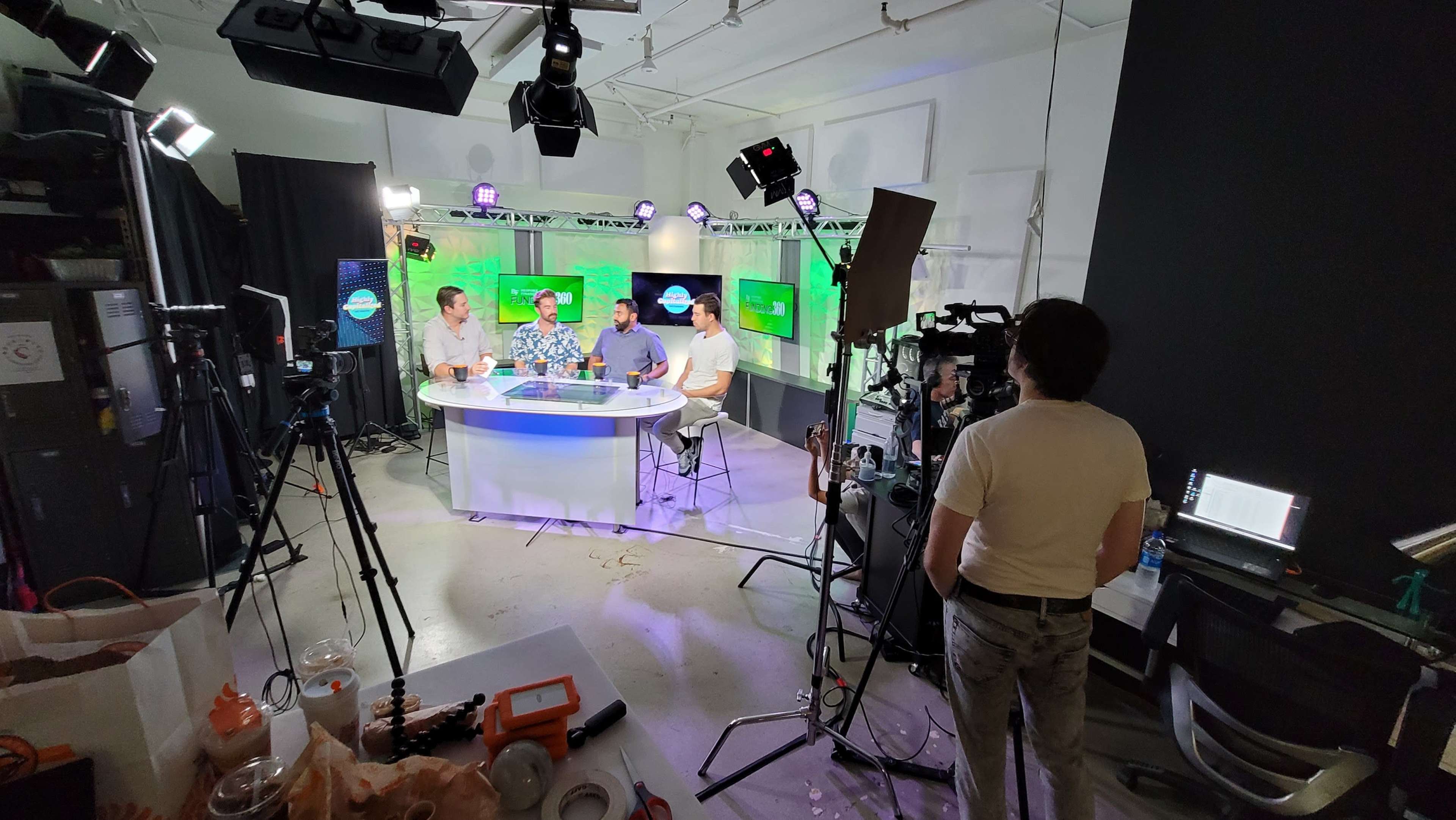 The image shows a television studio setup with four people seated at a table, surrounded by cameras and lights, while a green screen is visible in the background.