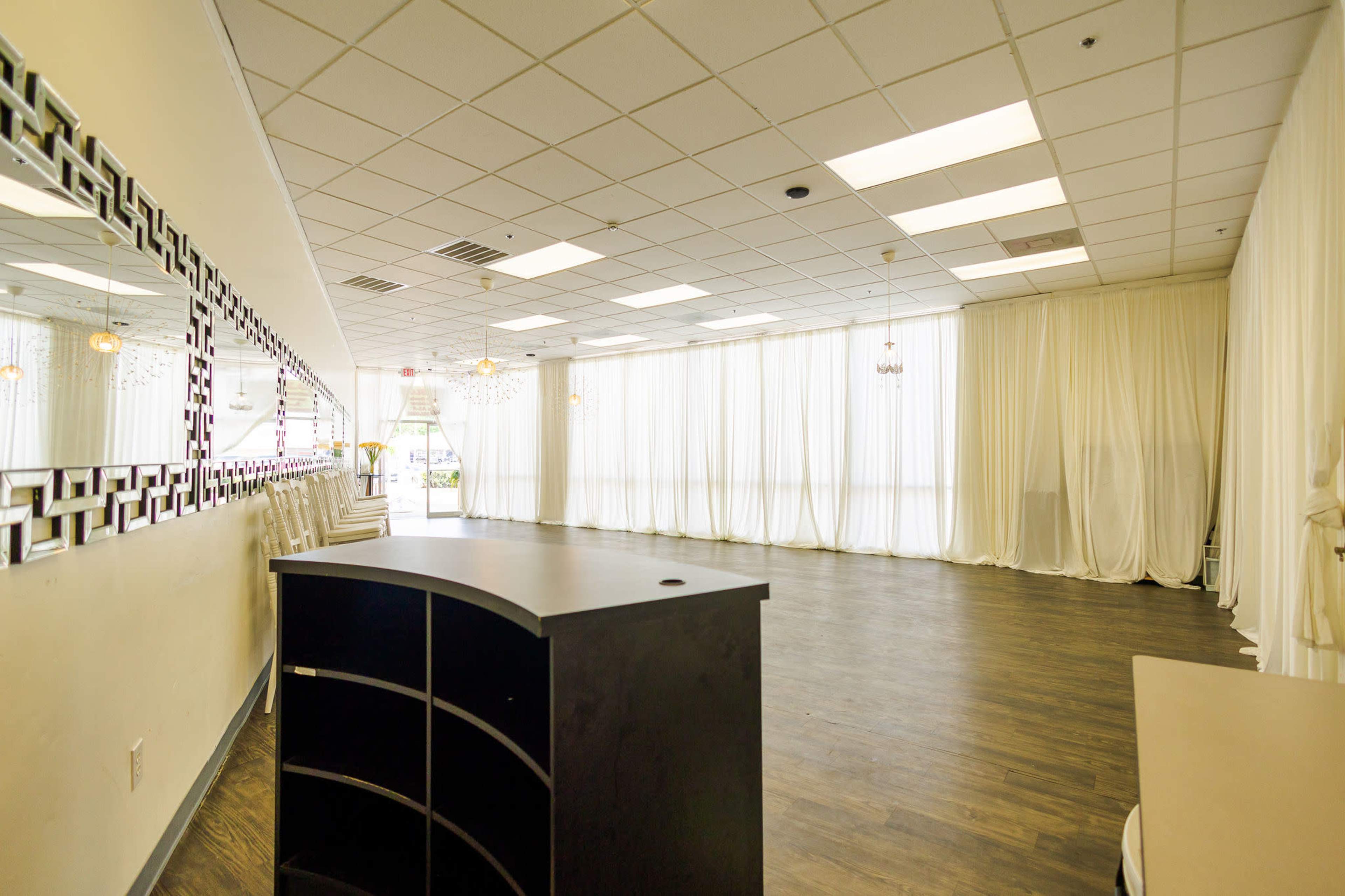 The image shows an empty event space with large windows covered by sheer curtains and a dark-colored reception desk in the foreground.