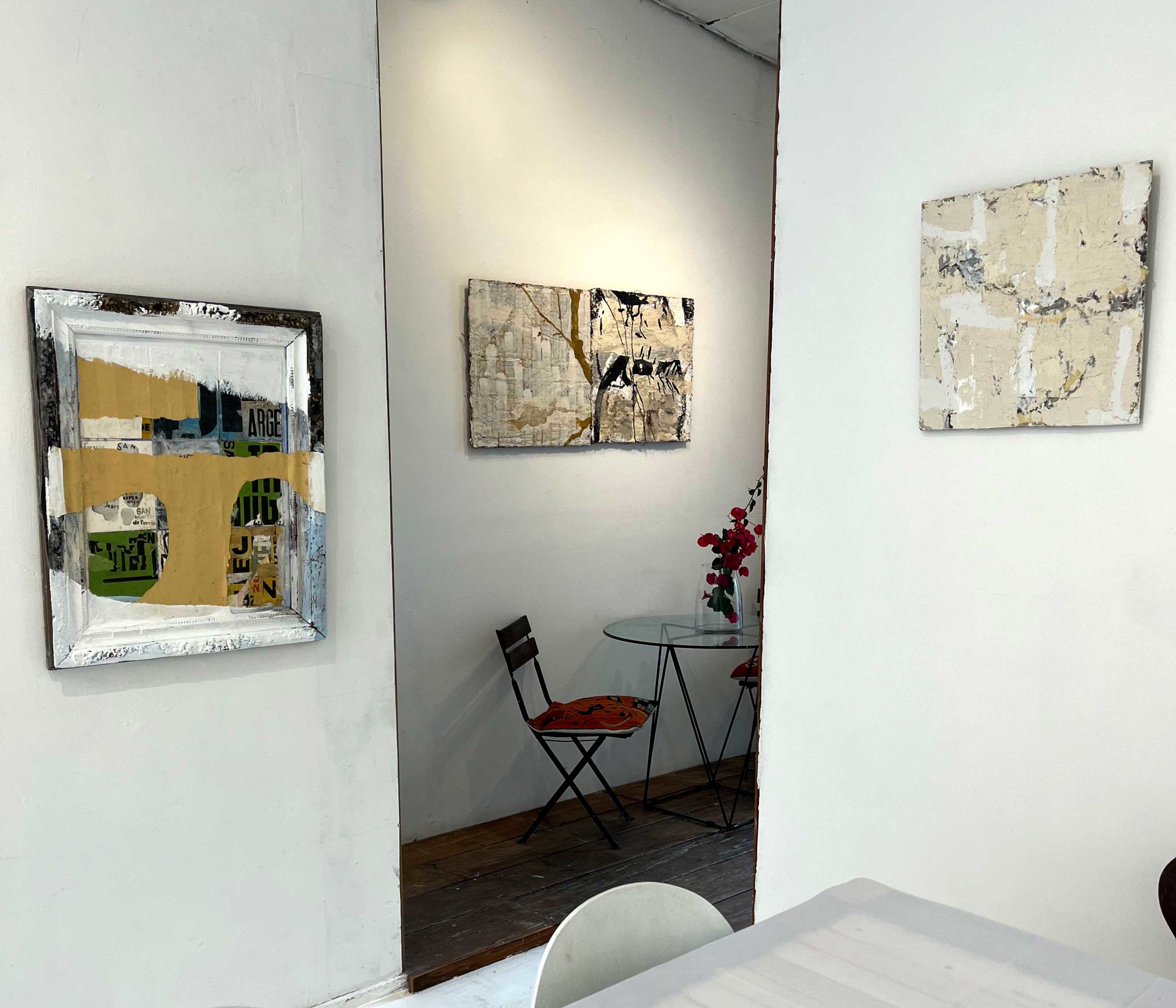 The image shows an artistic interior with three framed artworks on white walls, a small glass table with two chairs, and a vase of flowers on the table.