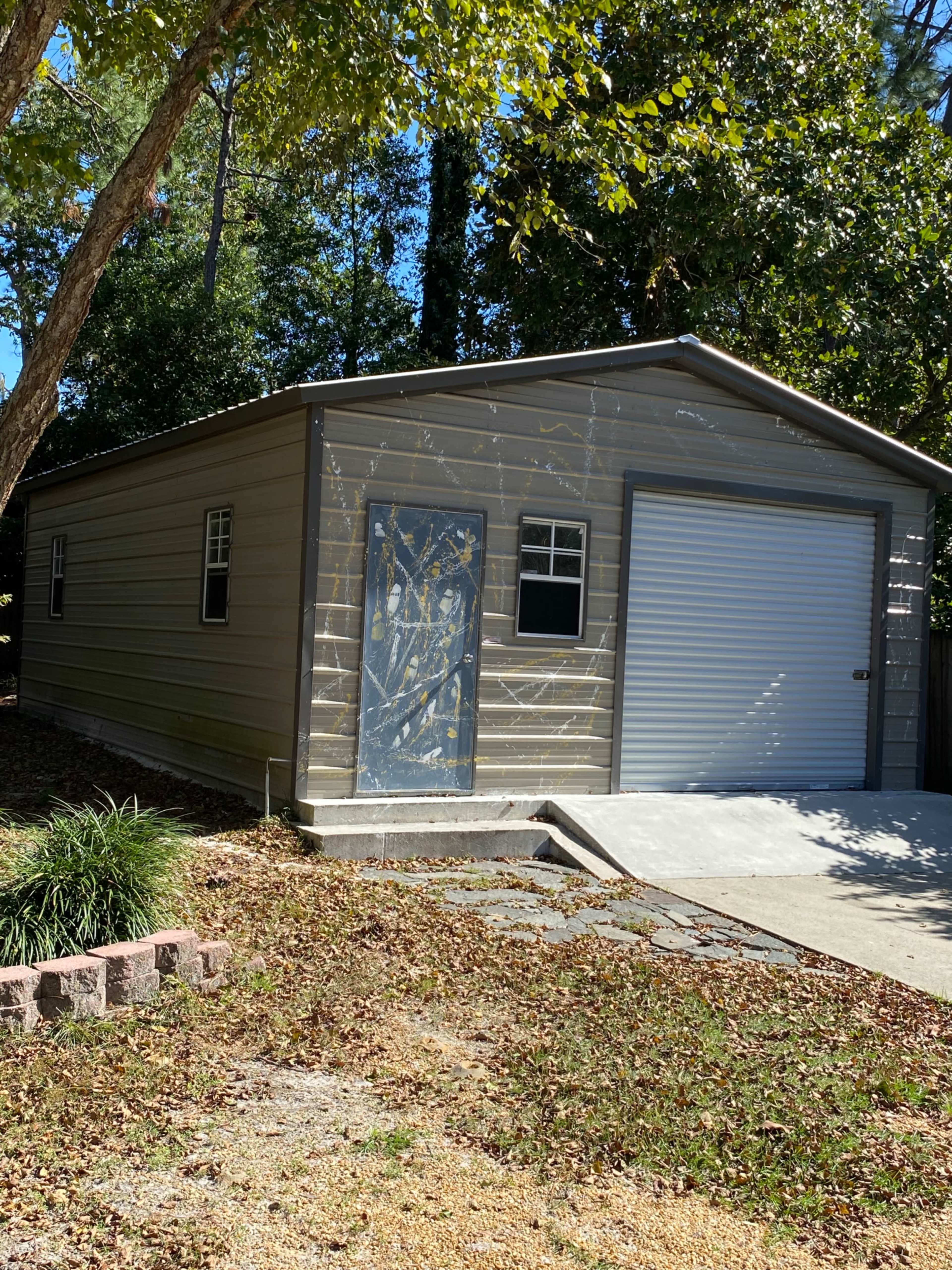 A metal storage shed with a roll-up door and a side window is situated on a gravel driveway surrounded by grass and trees.