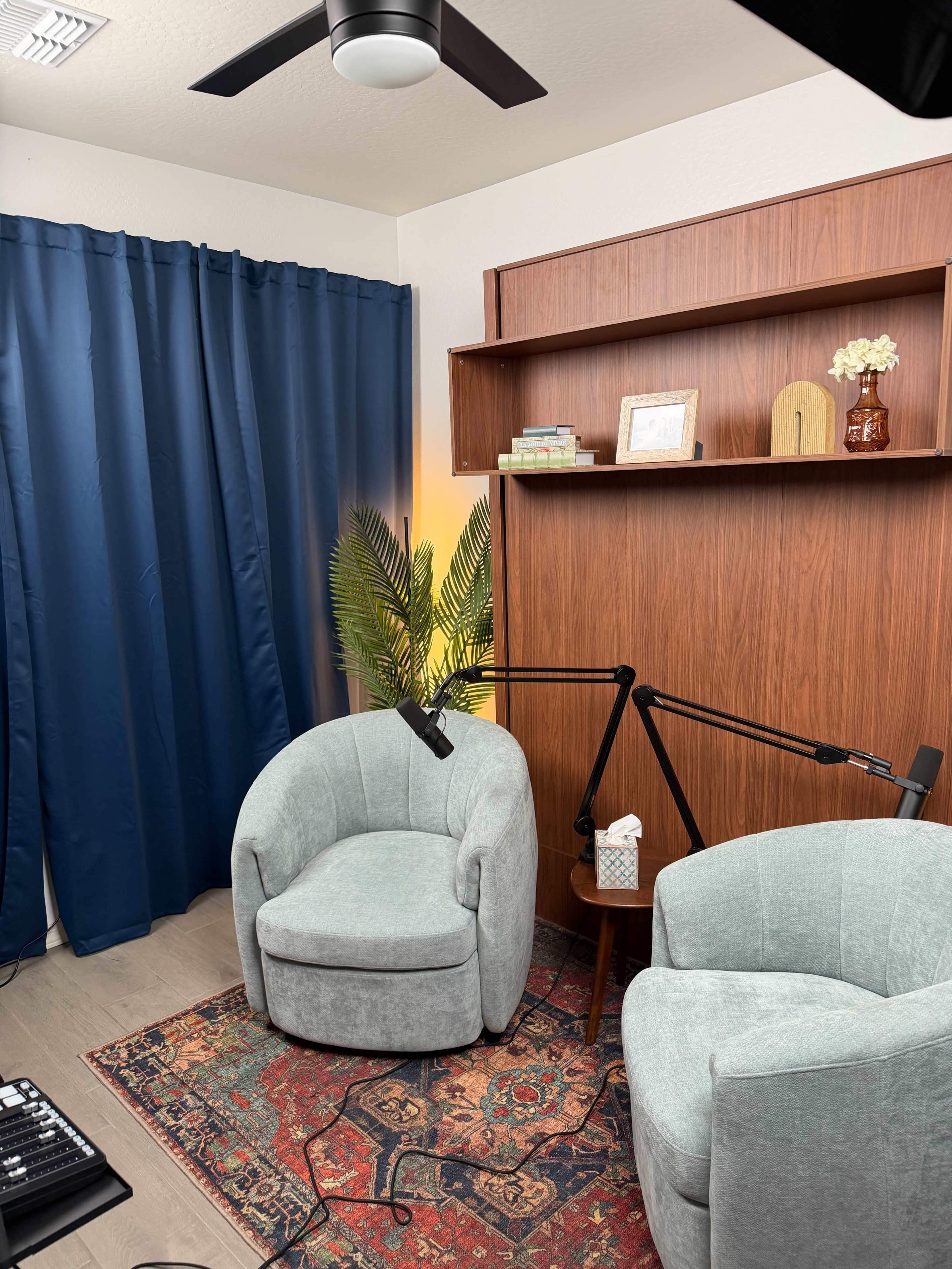 The image shows a cozy corner of a room with two upholstered chairs, a small round table, a shelf with decorative items, and a plant, all illuminated by soft lighting.