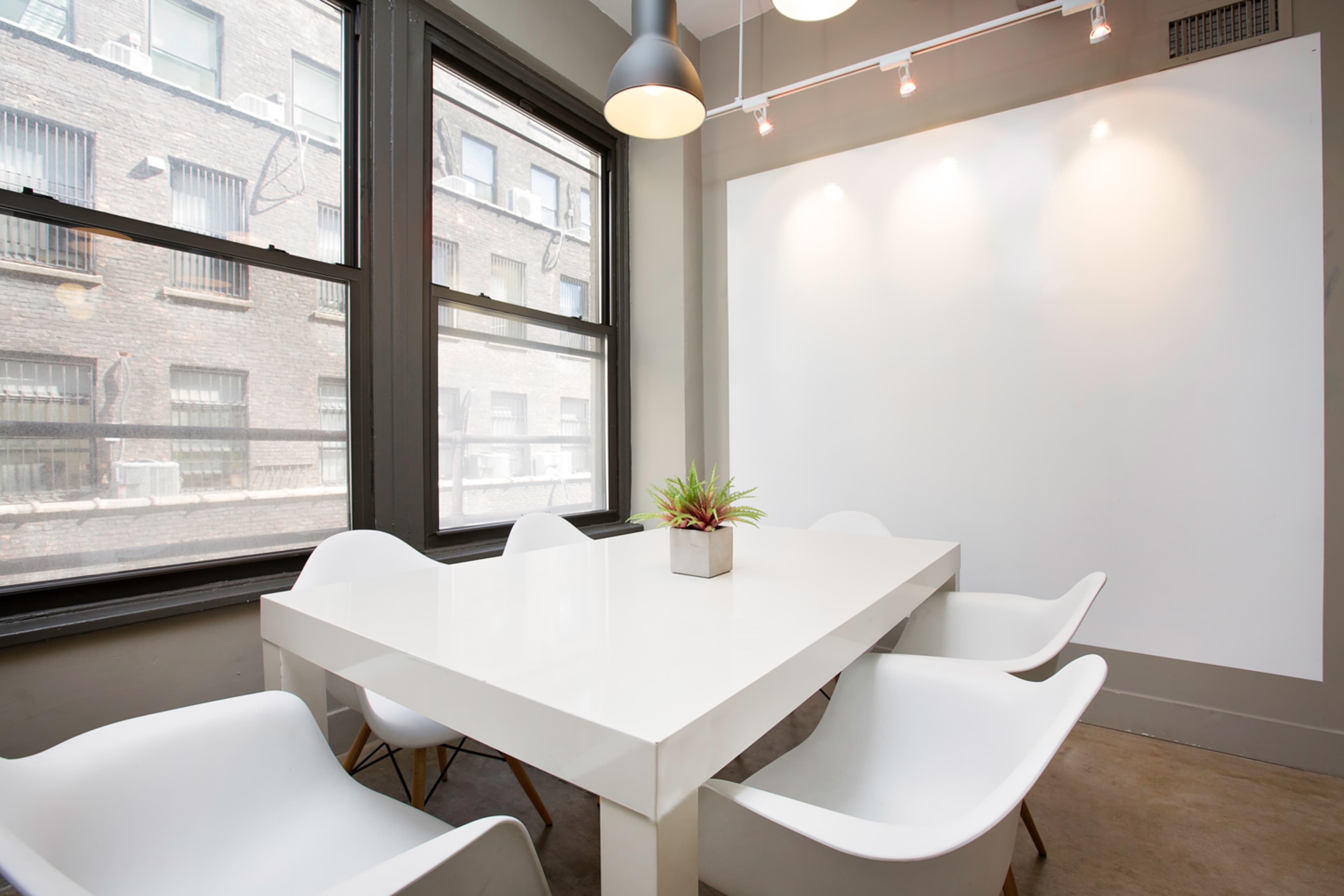 A modern conference room with a white table surrounded by four white chairs, illuminated by overhead lights and featuring large windows.