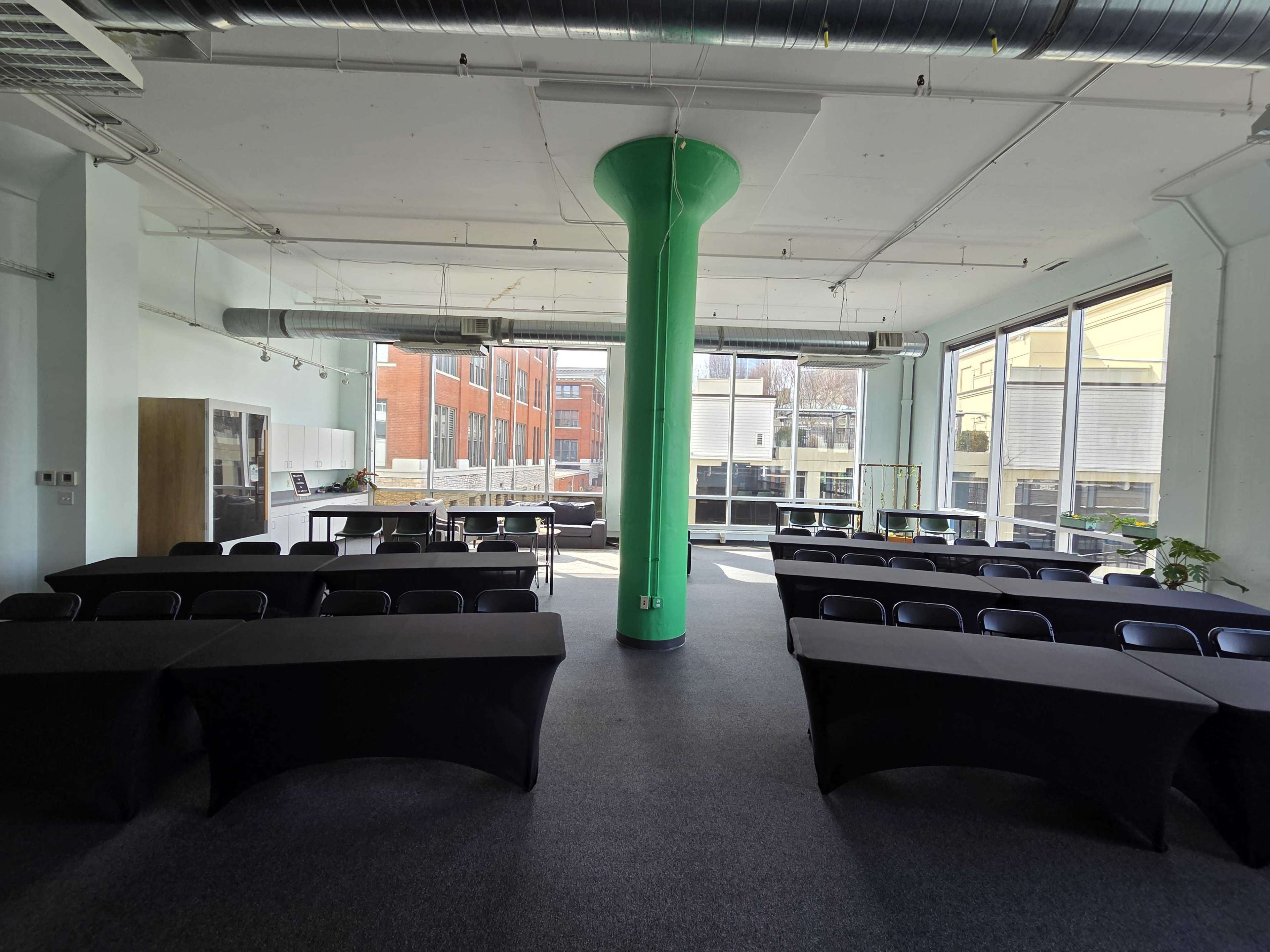 The image shows a spacious office room with several black tables arranged around a central green column and large windows allowing natural light in.