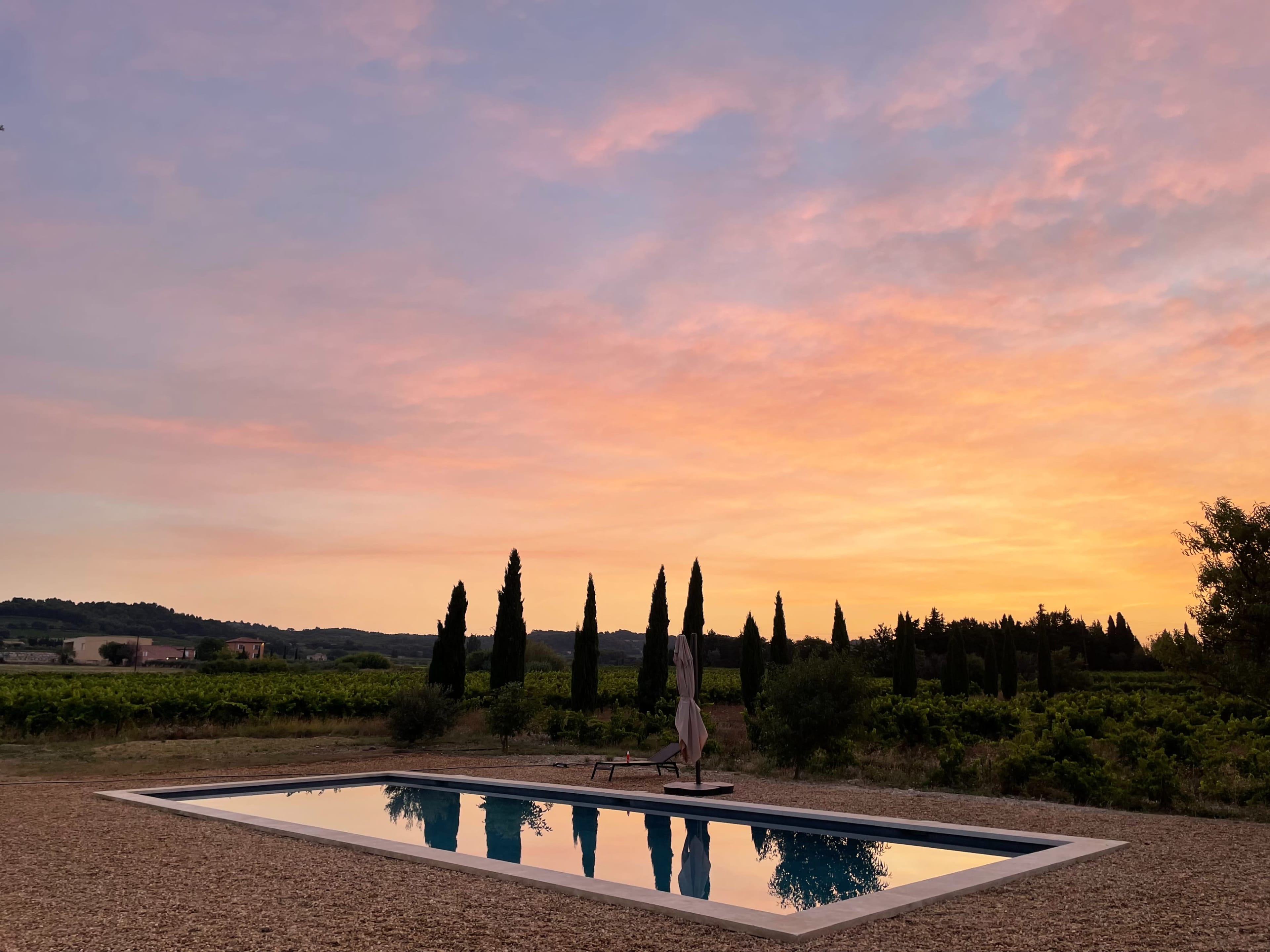 A swimming pool reflects a colorful sunset surrounded by cypress trees and vineyards.