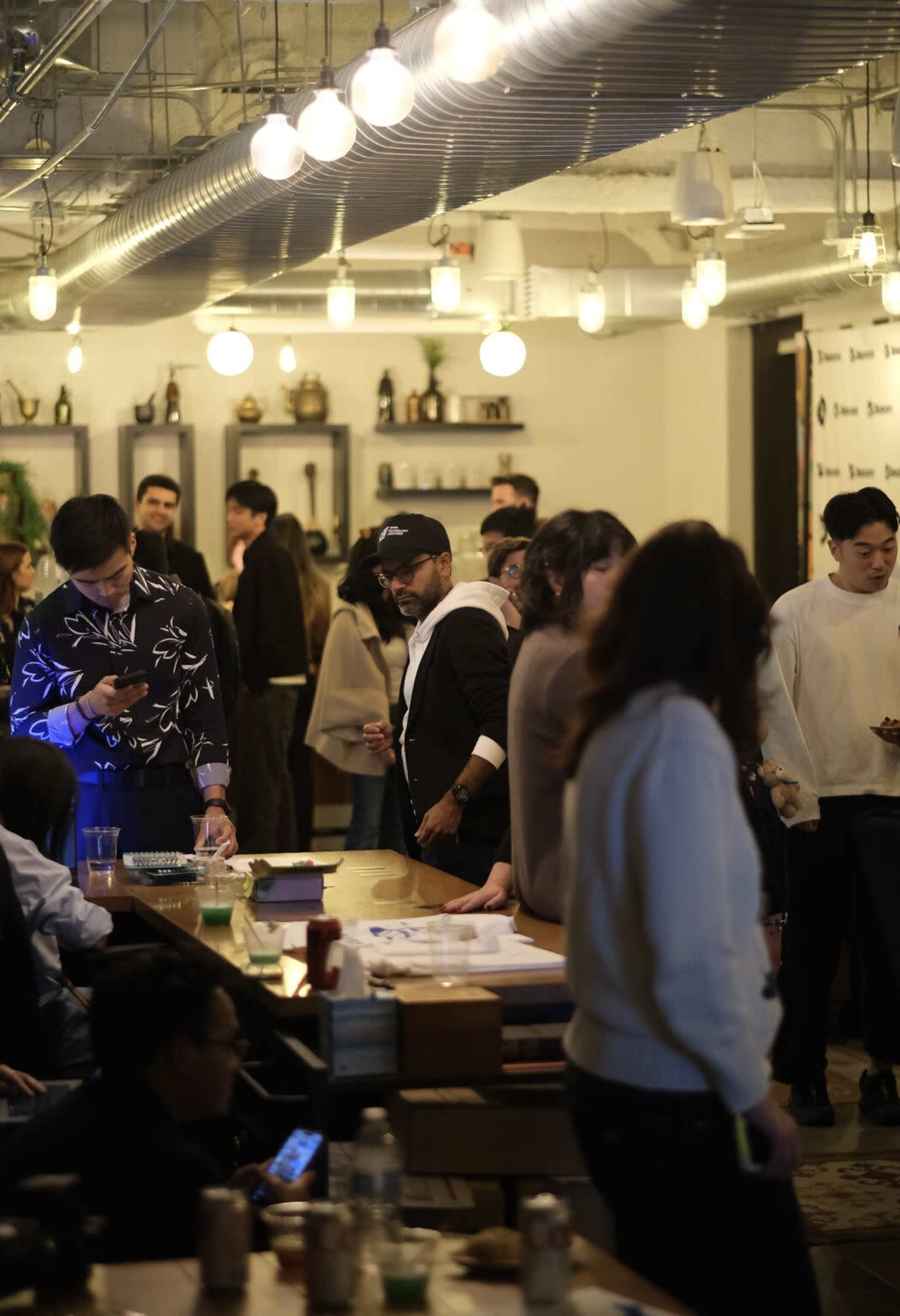 A busy social setting features people mingling and engaging in conversation, with tables set up in a well-lit indoor space.