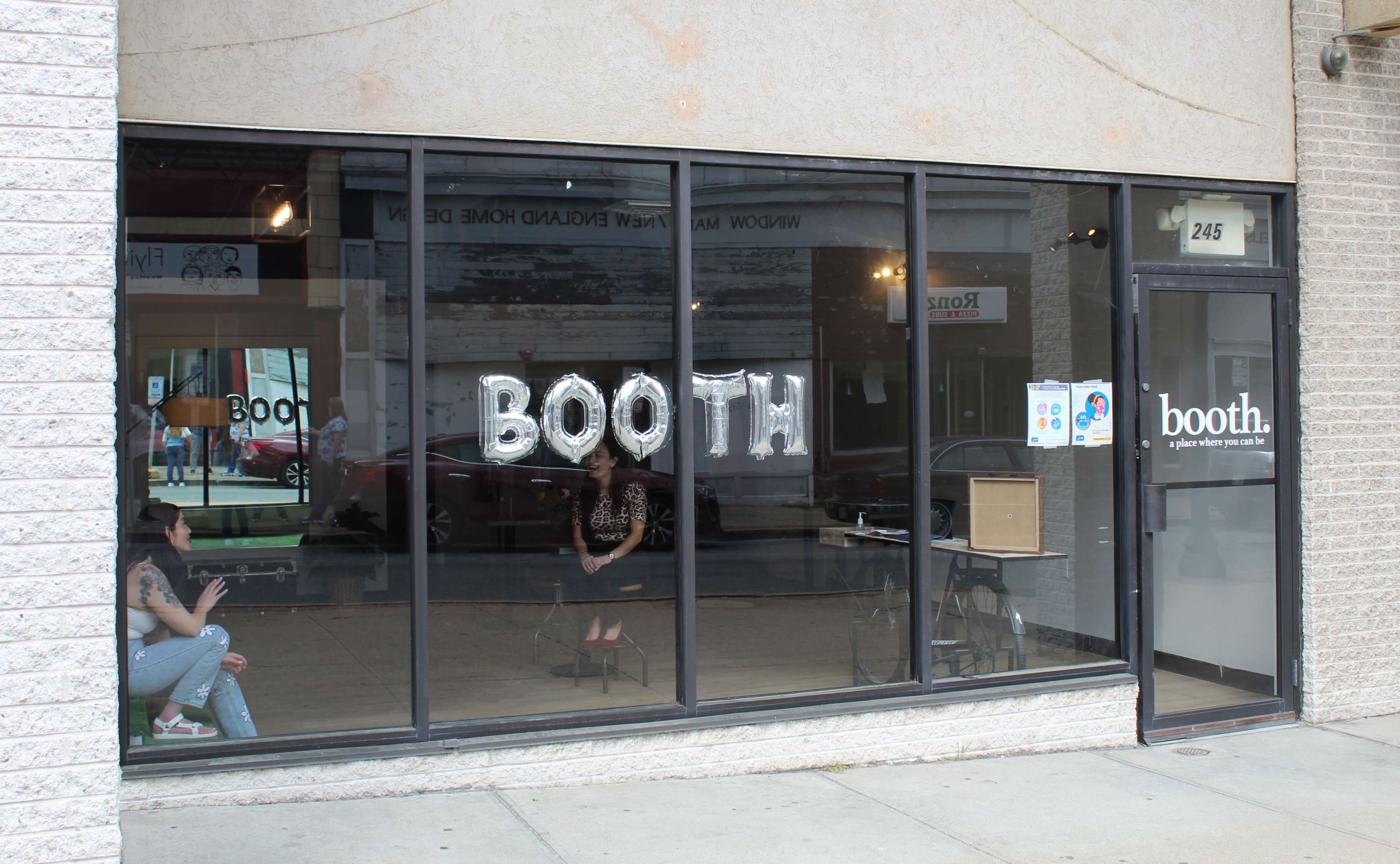 A storefront with large "BOOTH" letters made of balloons displayed in the windows, and two people sitting inside.