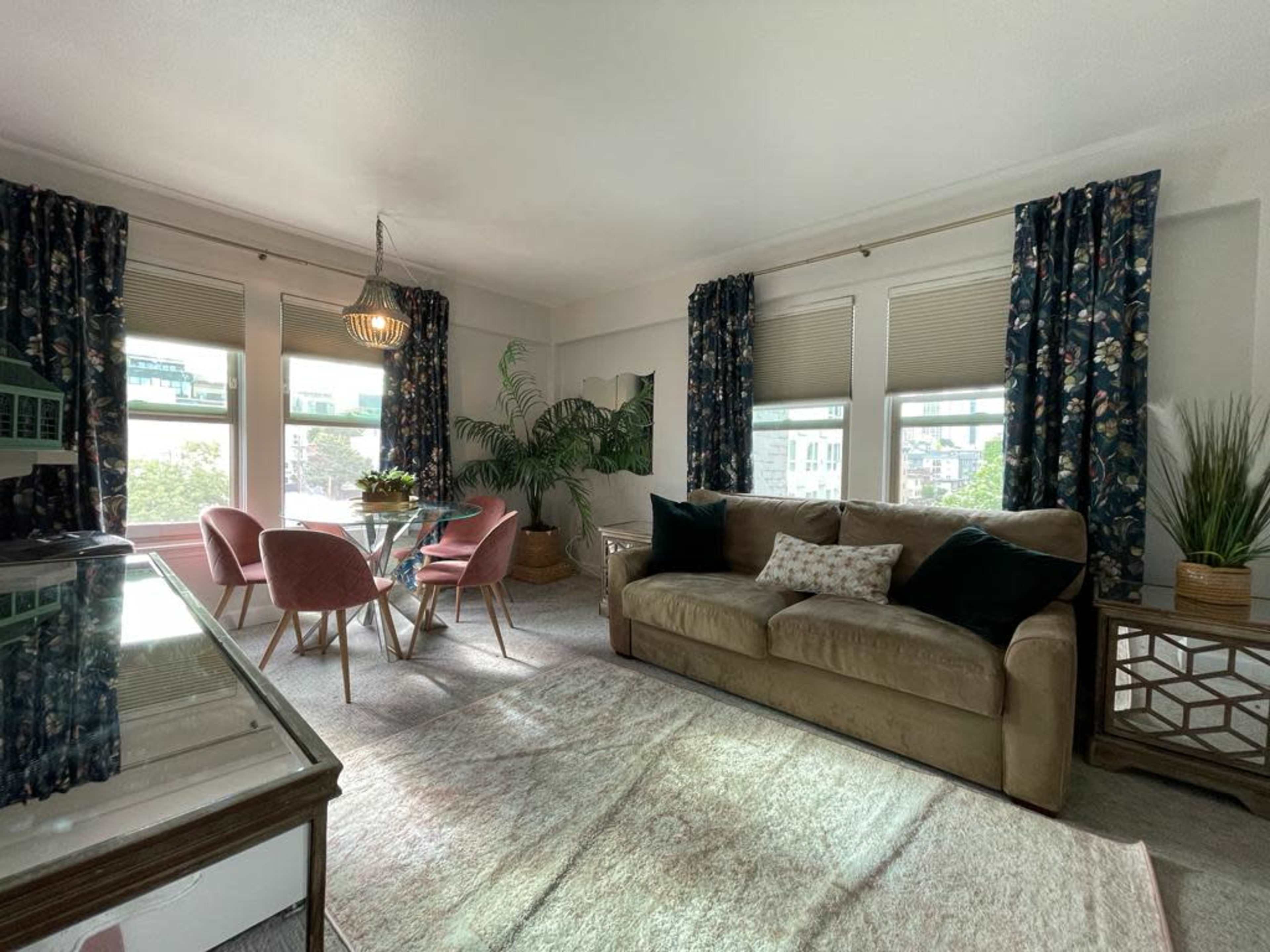The image shows a living room with a beige sofa, a glass dining table surrounded by pink chairs, large windows with patterned curtains, and indoor plants.