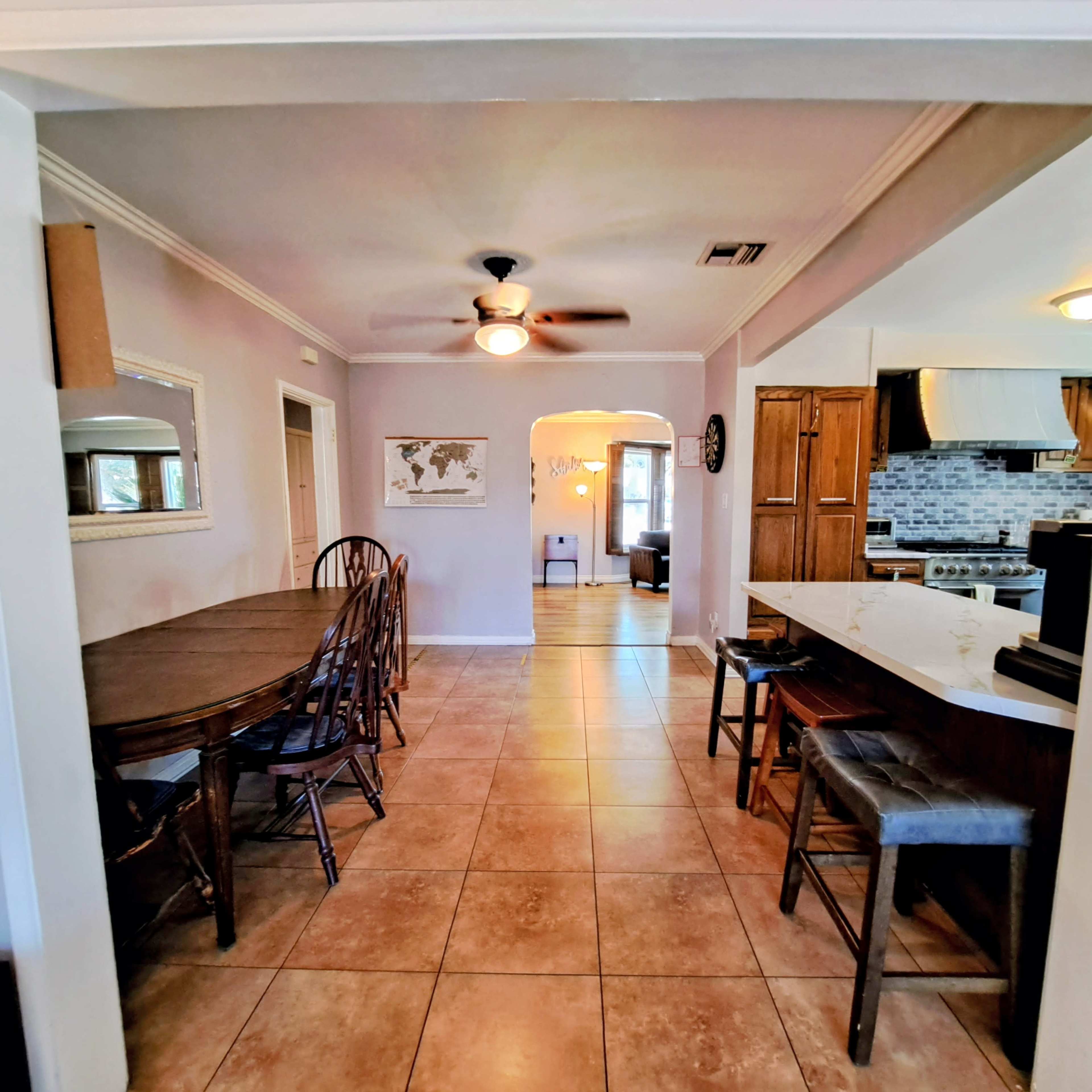 The image shows a spacious dining area with a long wooden table and chairs, leading to a kitchen featuring a bar counter with stools and tile flooring.