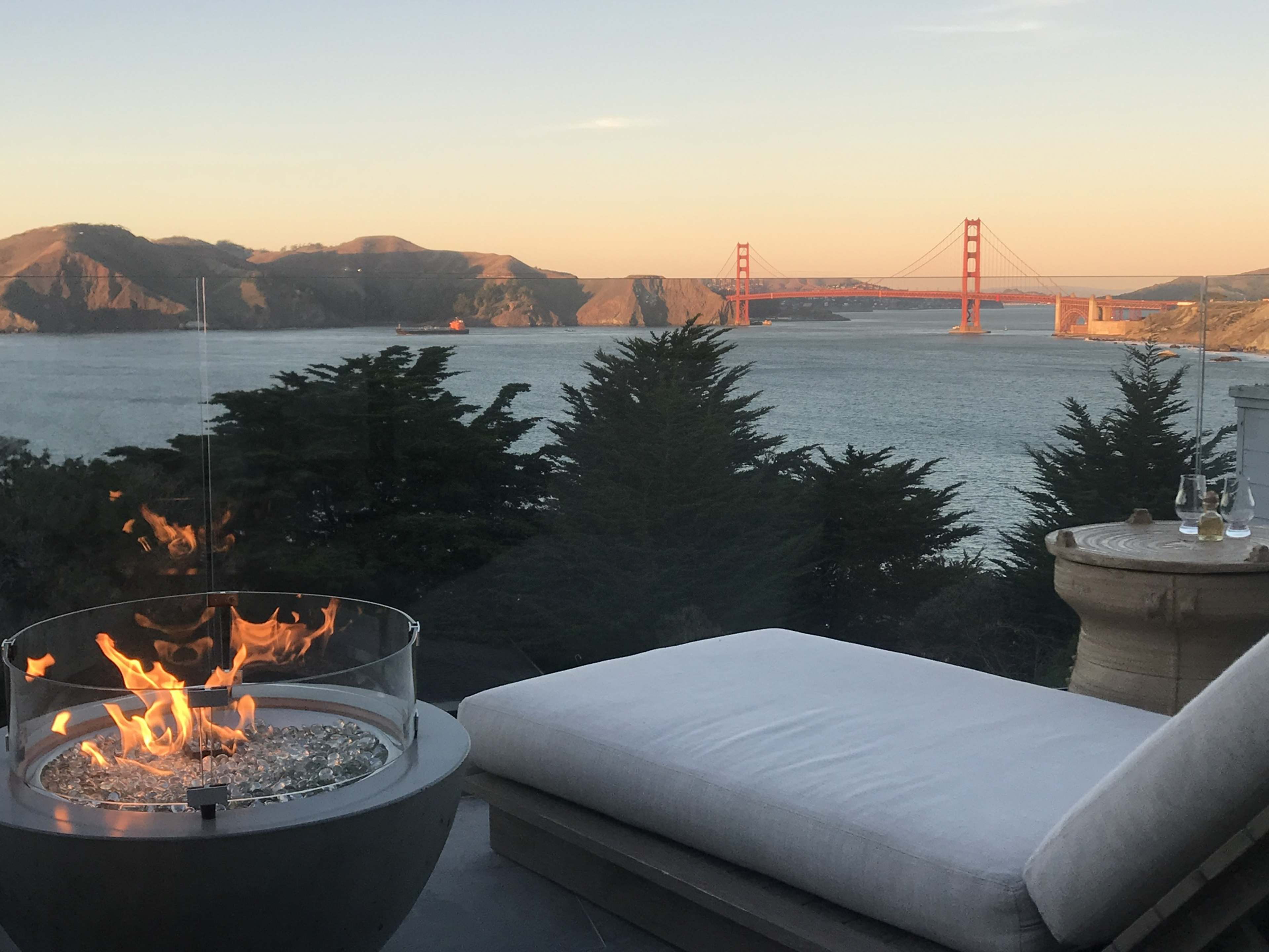 A modern seating area features a circular fire pit with flames, overlooking the Golden Gate Bridge and surrounding hills at sunset.