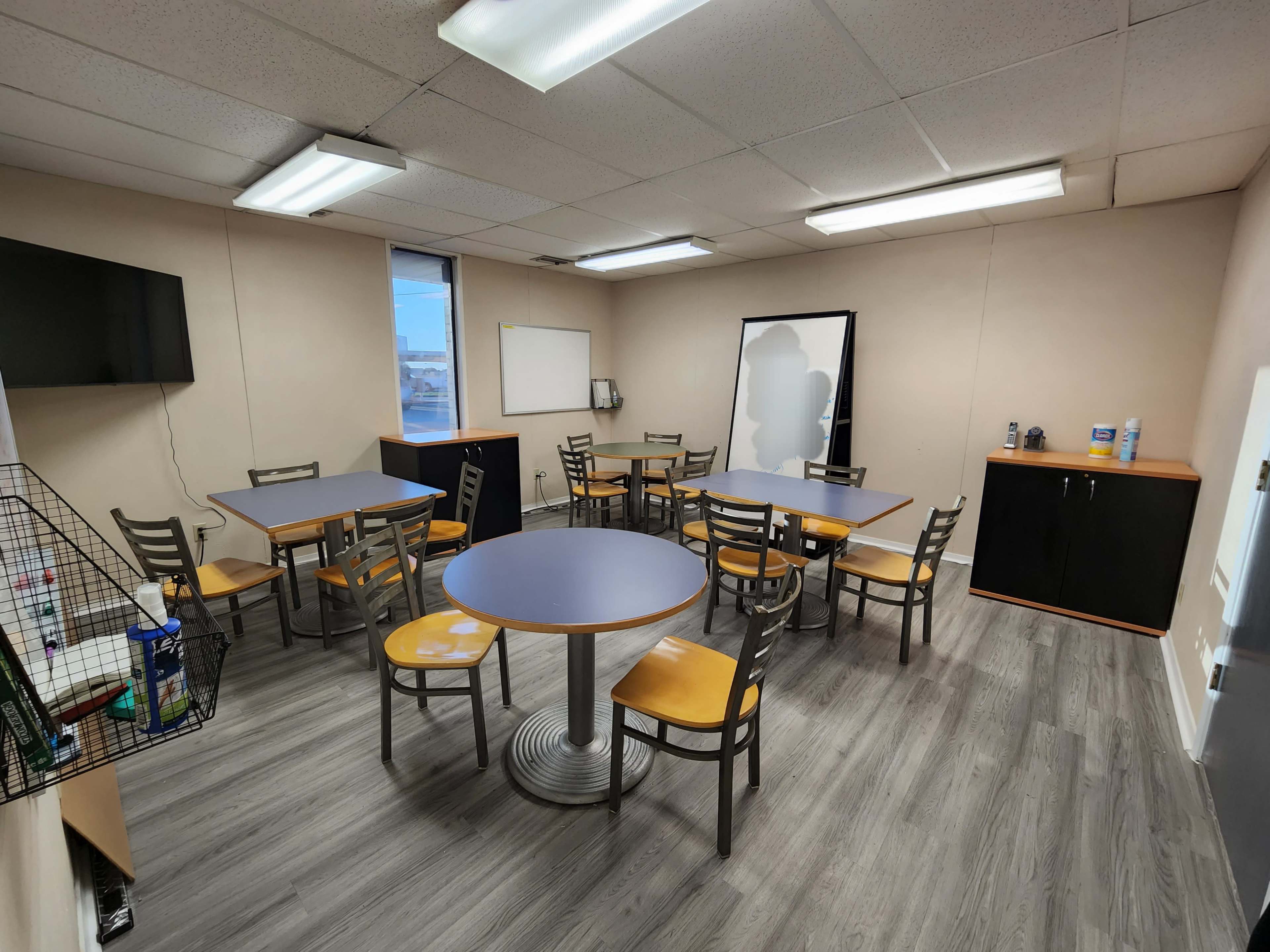 A well-lit room with multiple tables and chairs arranged for collaborative seating, featuring a TV, a whiteboard, and storage cabinets.