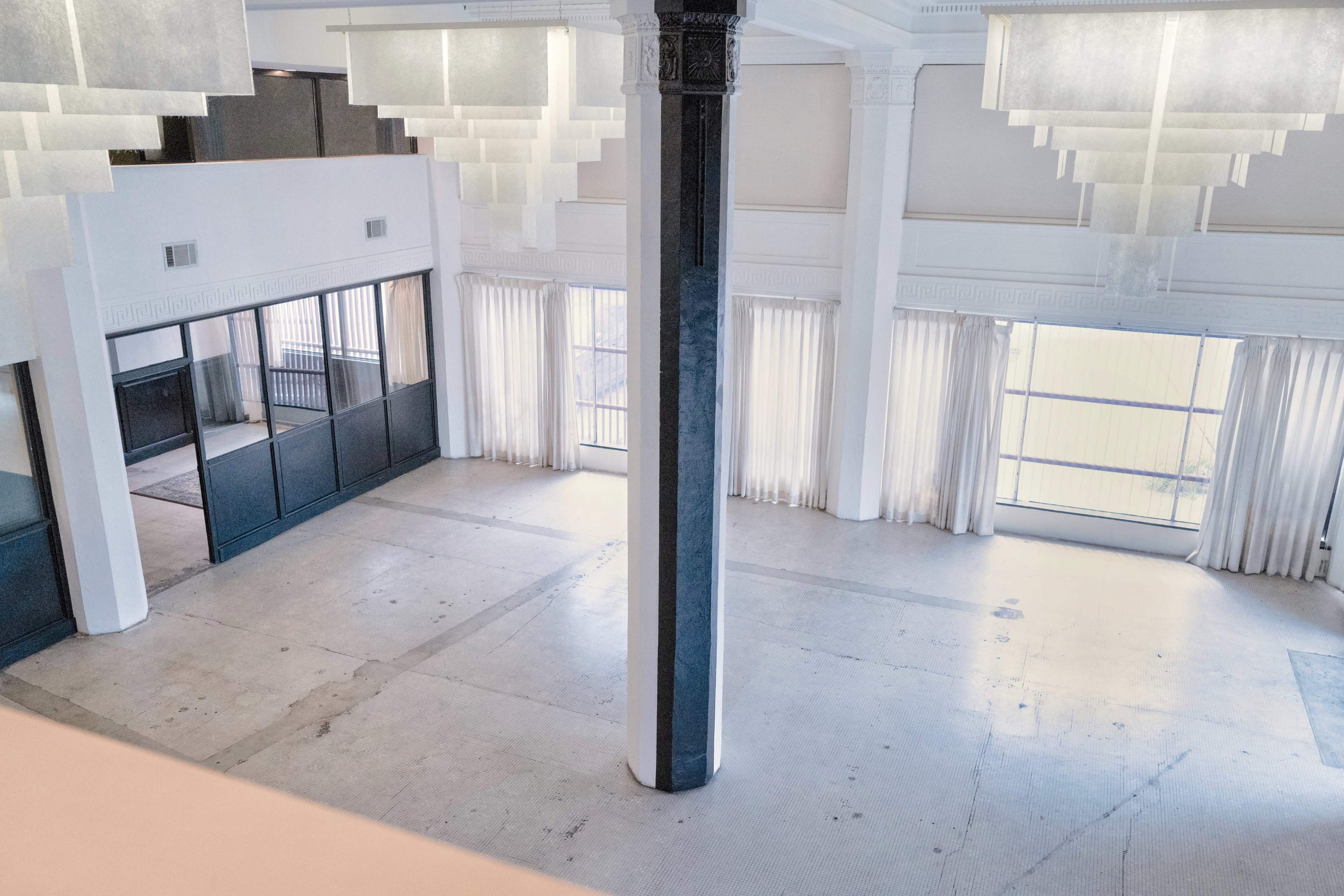 The image shows a spacious, bright room with large windows, white curtains, and a central column, featuring minimal furnishings and a polished concrete floor.
