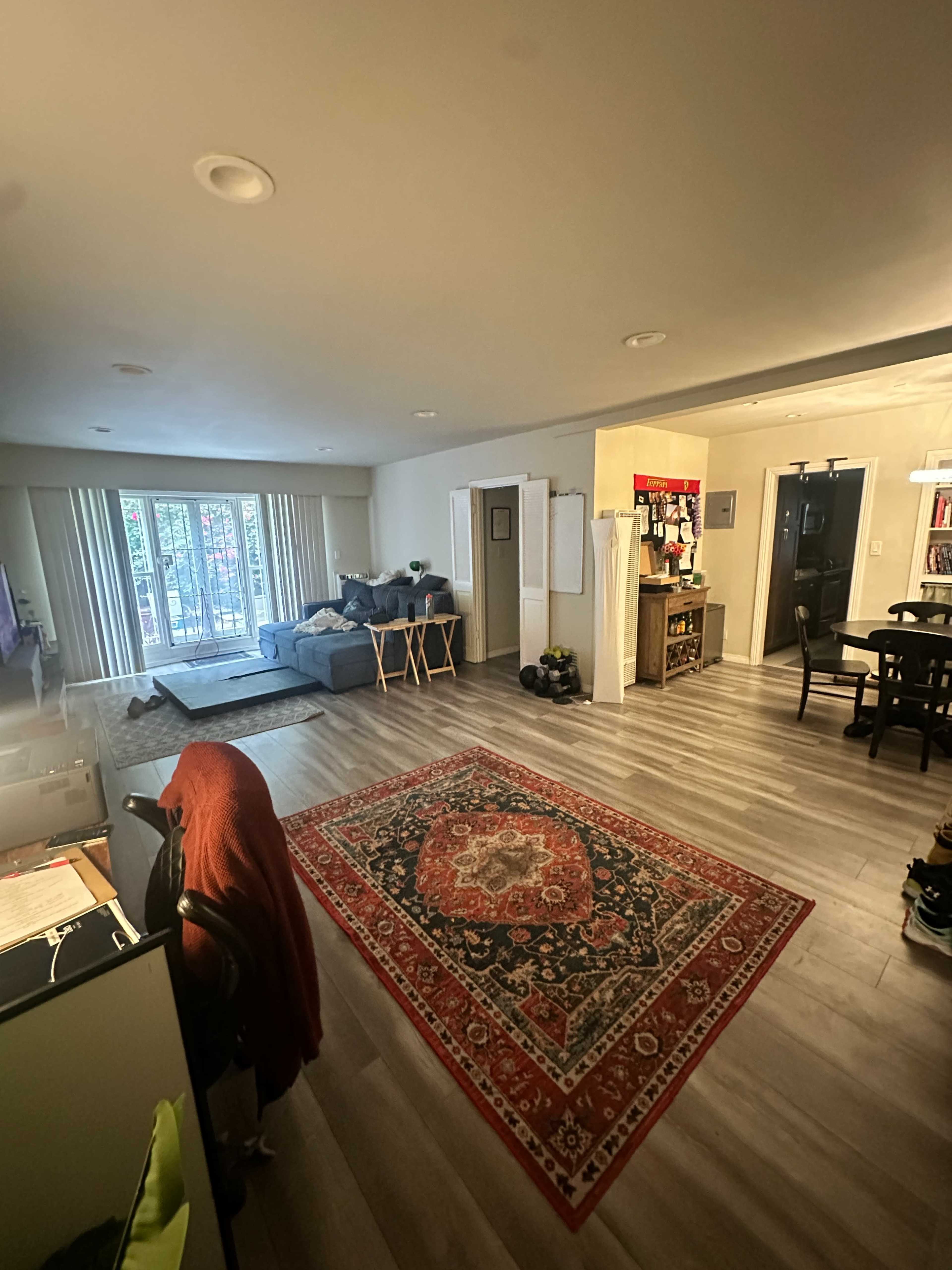 The image shows a well-lit living room with a sofa, dining area, and hardwood flooring, featuring a decorative rug in the center.