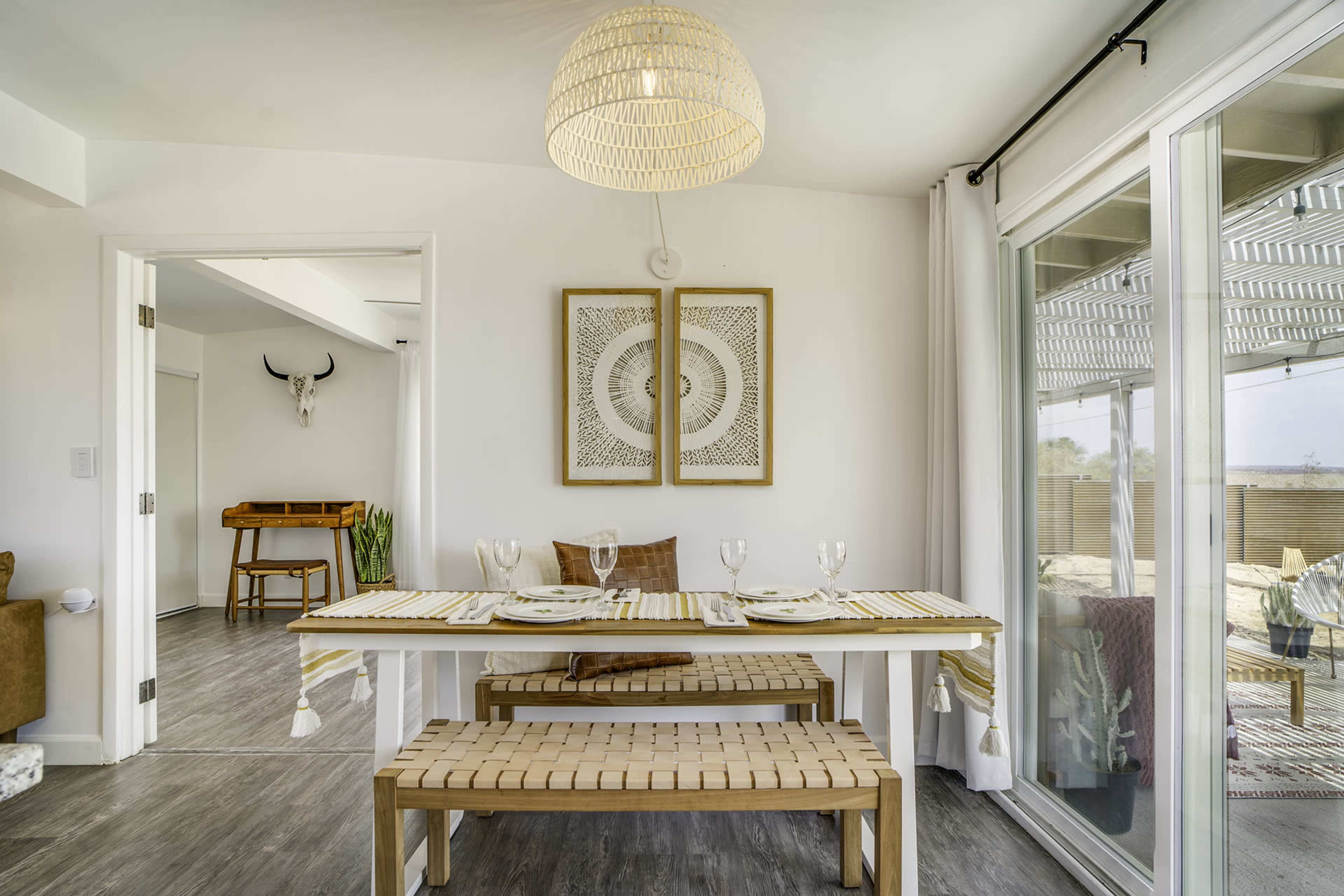 A dining area features a rectangular table set for four, surrounded by wooden benches, with a large light fixture overhead and a decorative wall piece in the background.