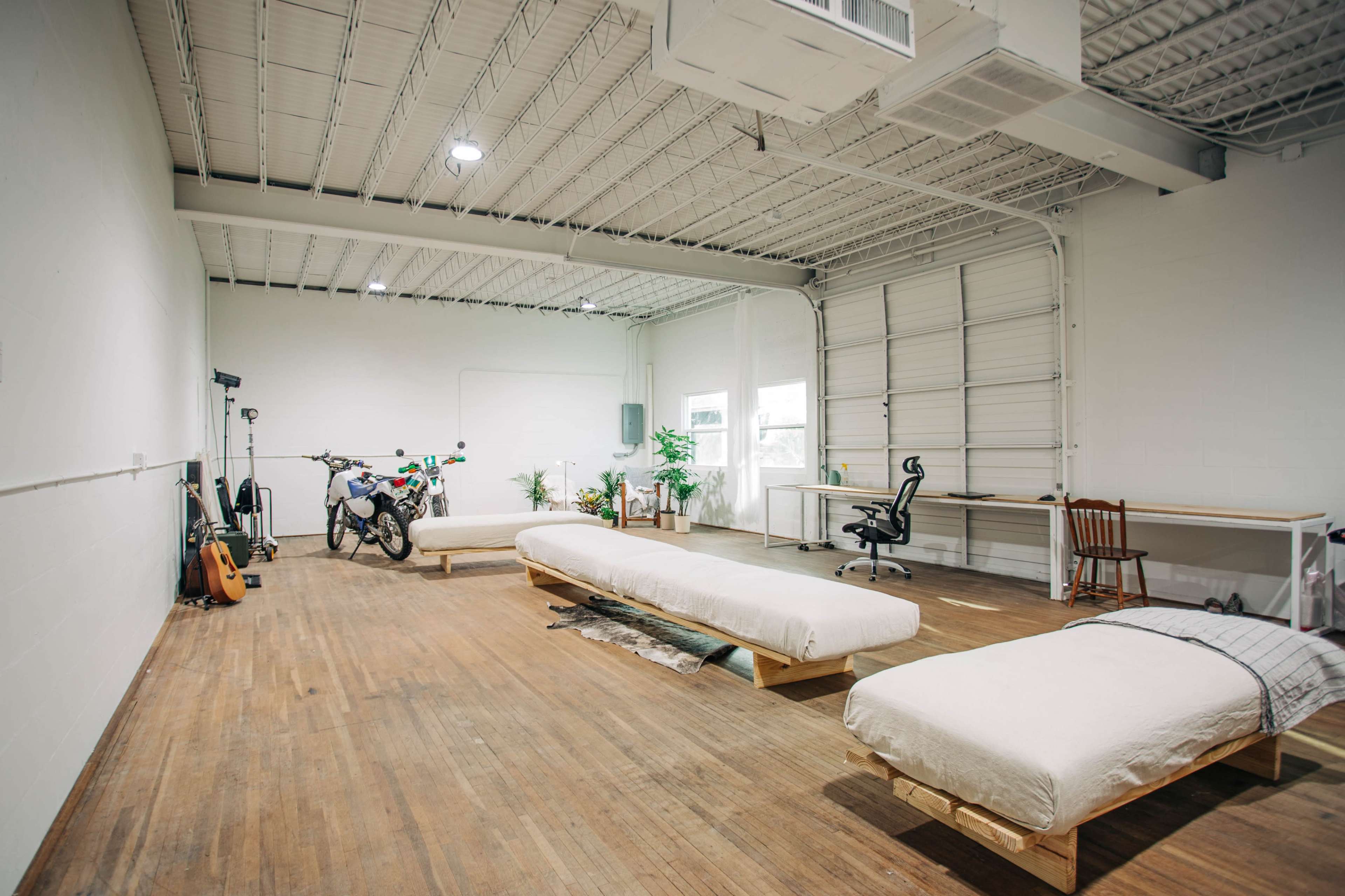 A spacious, minimalist room with wooden floors, two simple beds on wooden frames, a chair, a desk, a bicycle, and some plants.