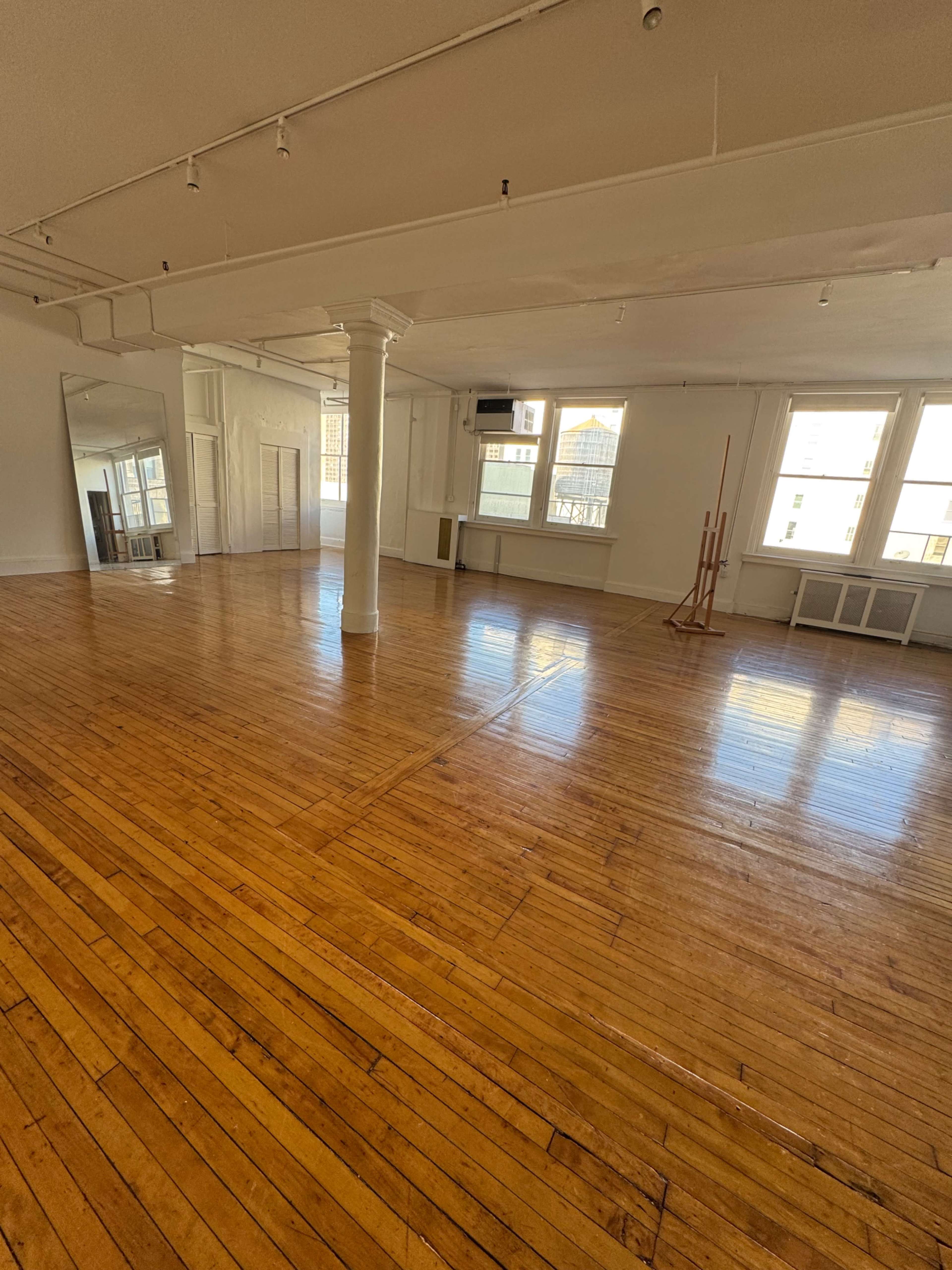 The image shows a spacious, empty room with polished wooden floors, large windows, and white walls.