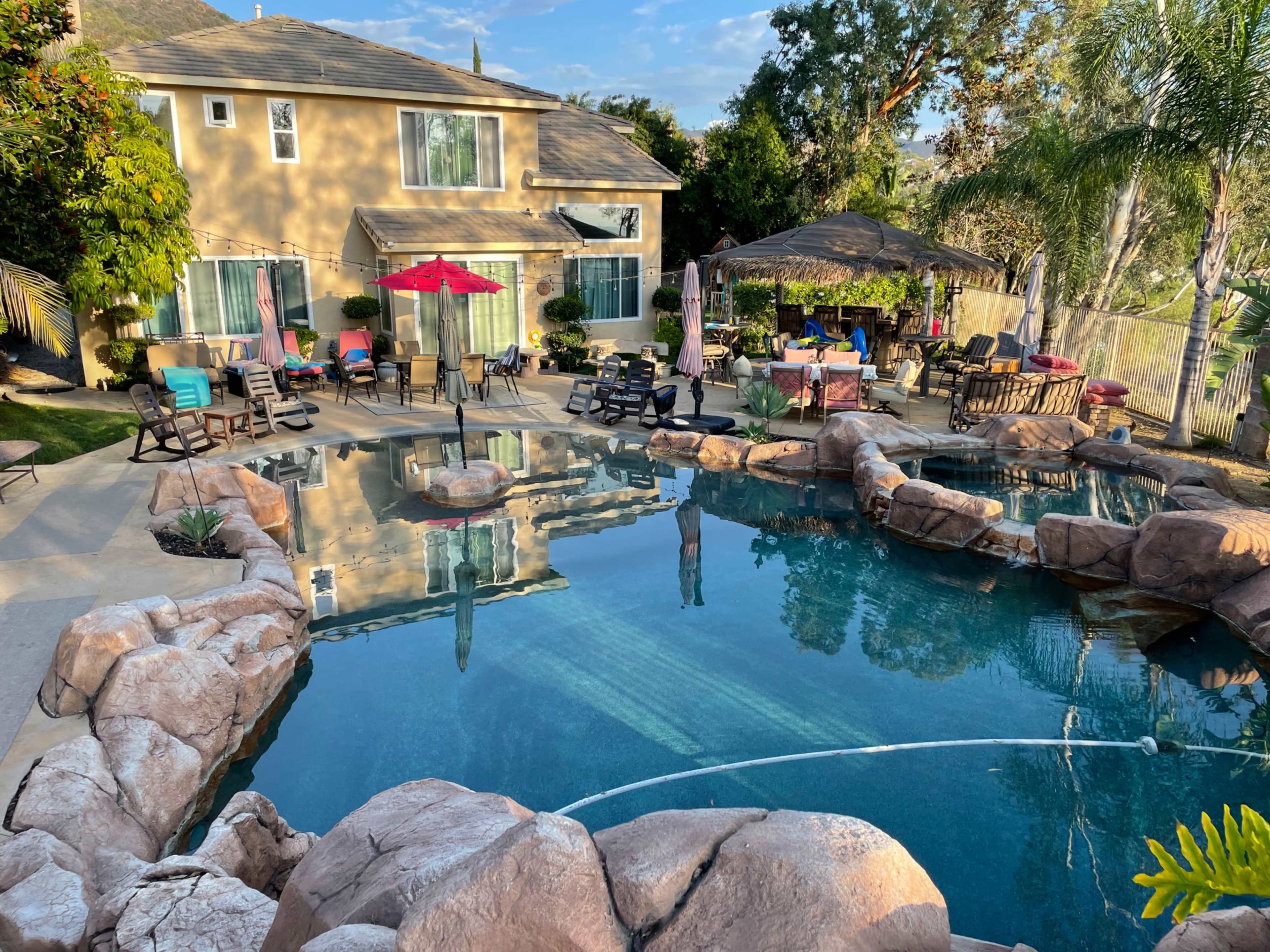 A backyard features a swimming pool surrounded by rock formations, lounge chairs, and a gazebo, with a two-story house in the background.