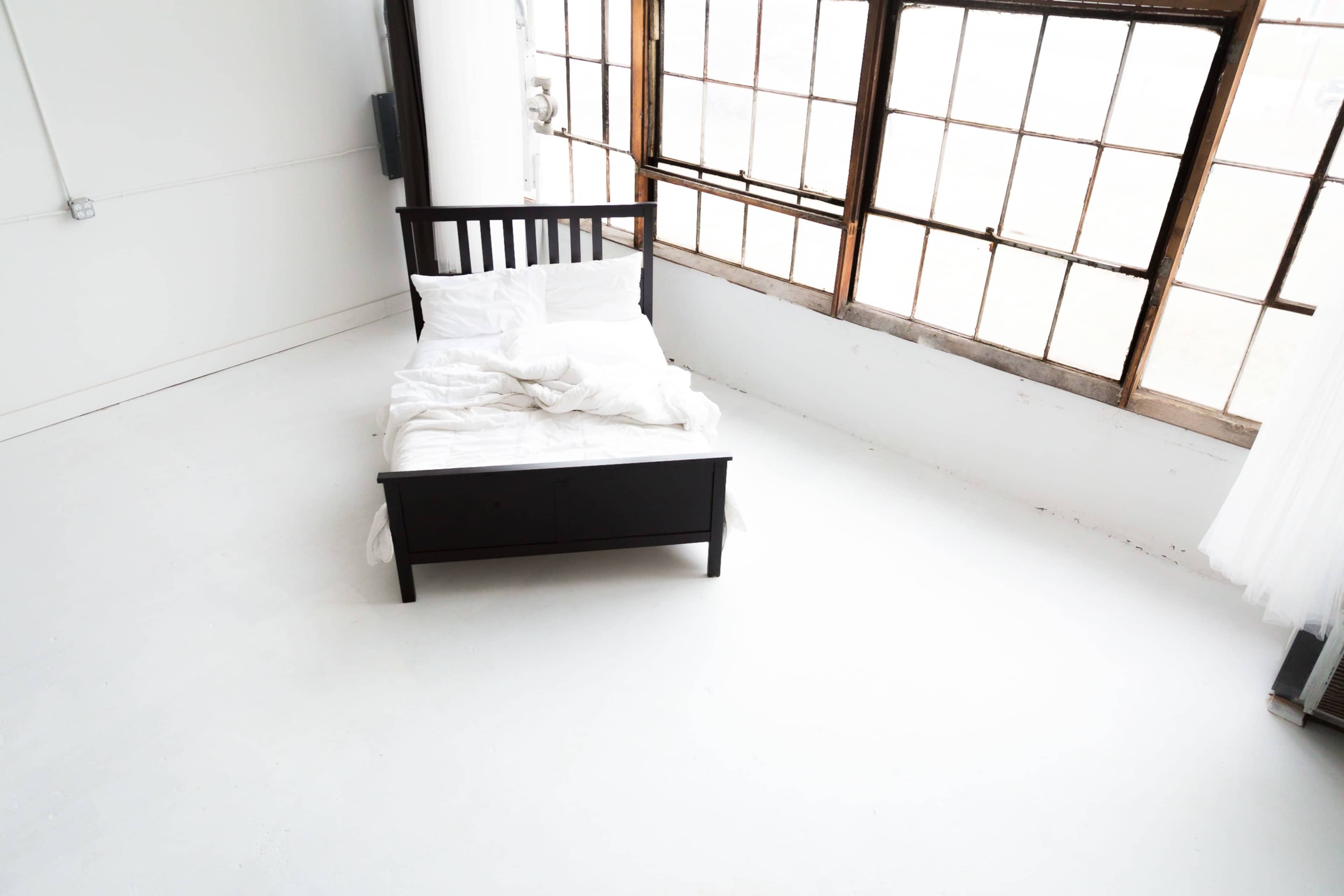 A black bed with white bedding is positioned in a large, sunlit room with tall windows.