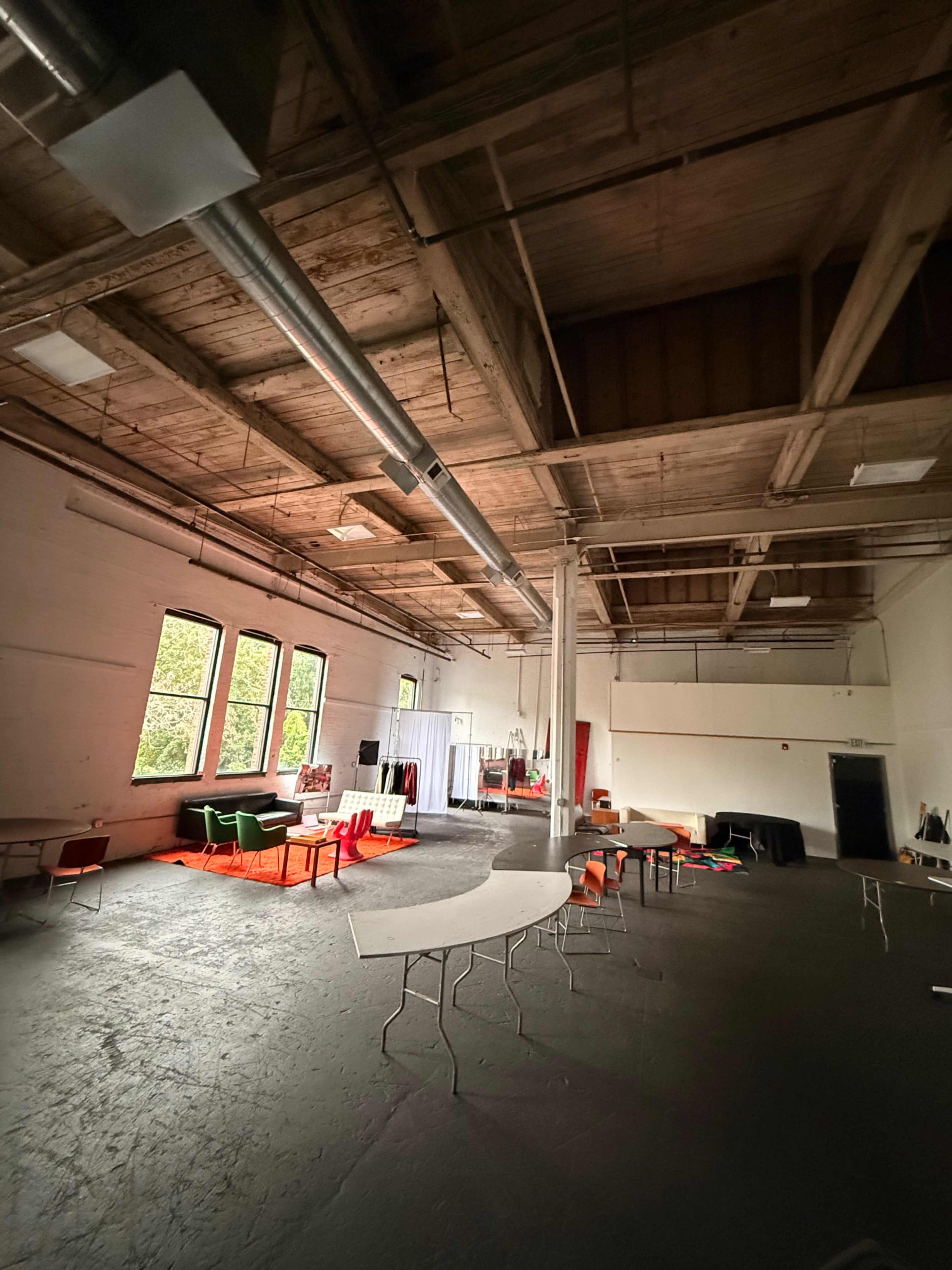 The image shows a spacious, empty room with wooden beams, large windows, and various tables and chairs scattered throughout.