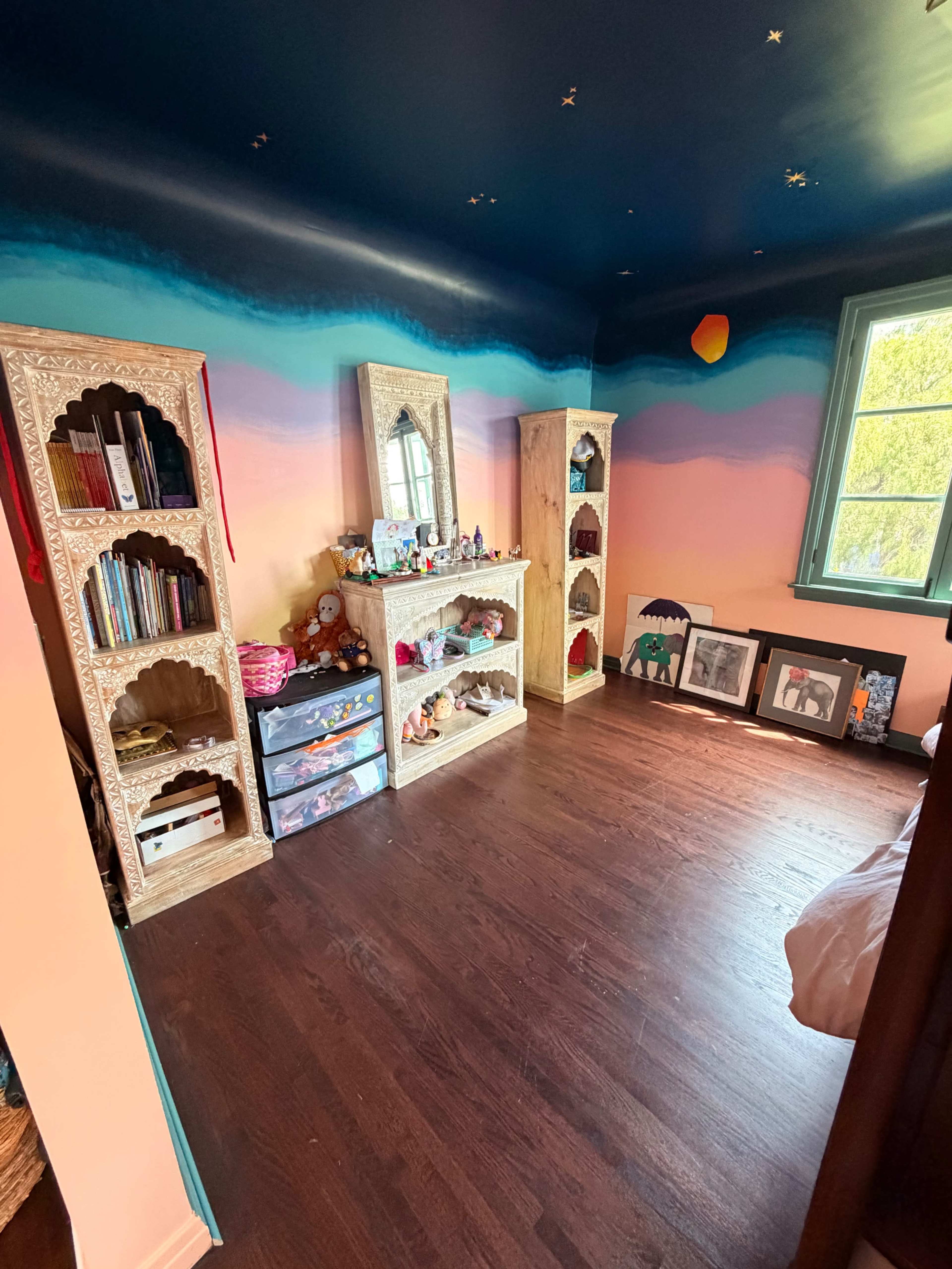 The image shows a brightly colored room with two ornate shelves filled with books and toys, and a large window allowing natural light to enter.
