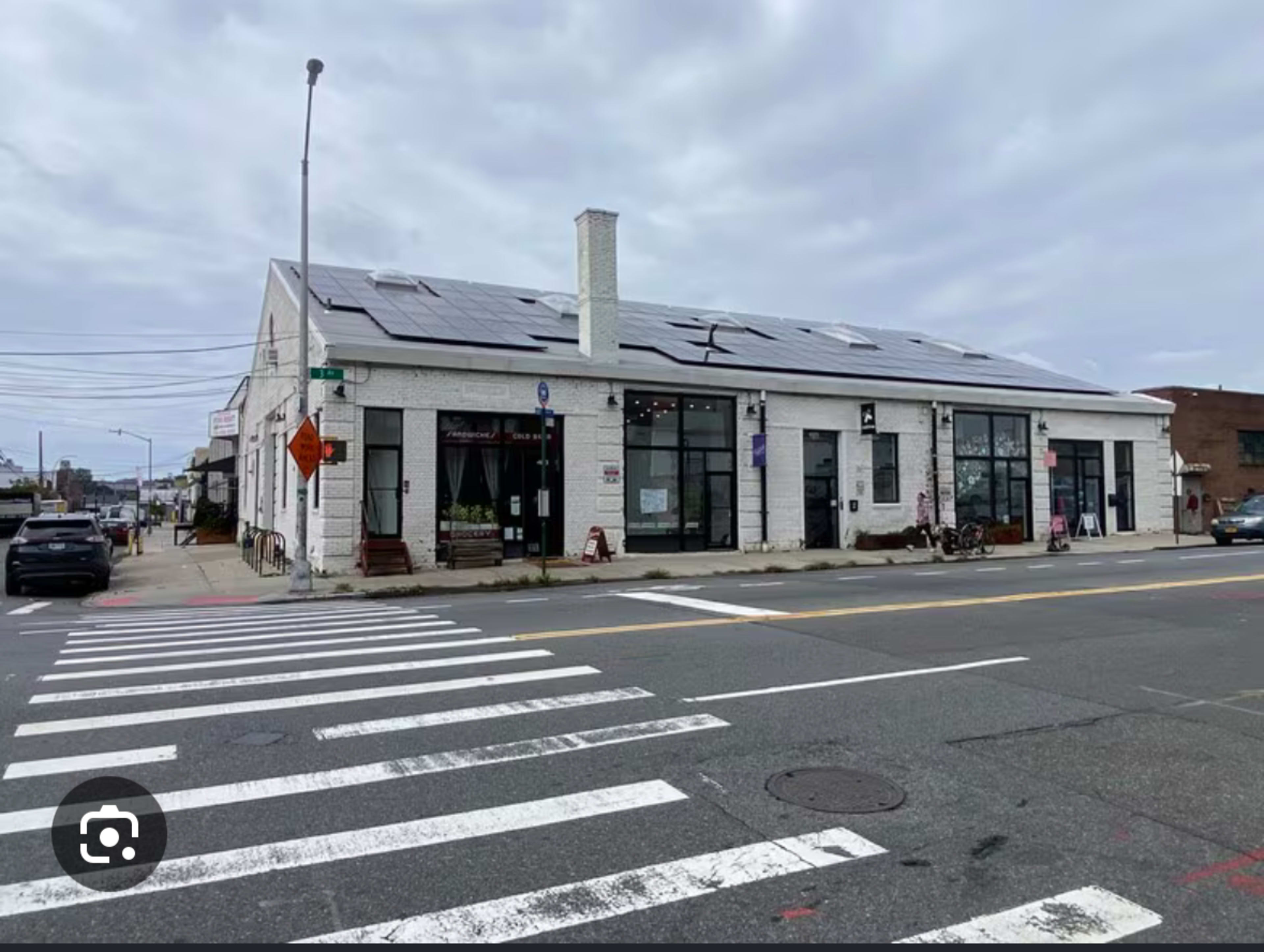 The image shows a large, white building with solar panels on the roof, located at an intersection with a crosswalk in the foreground.