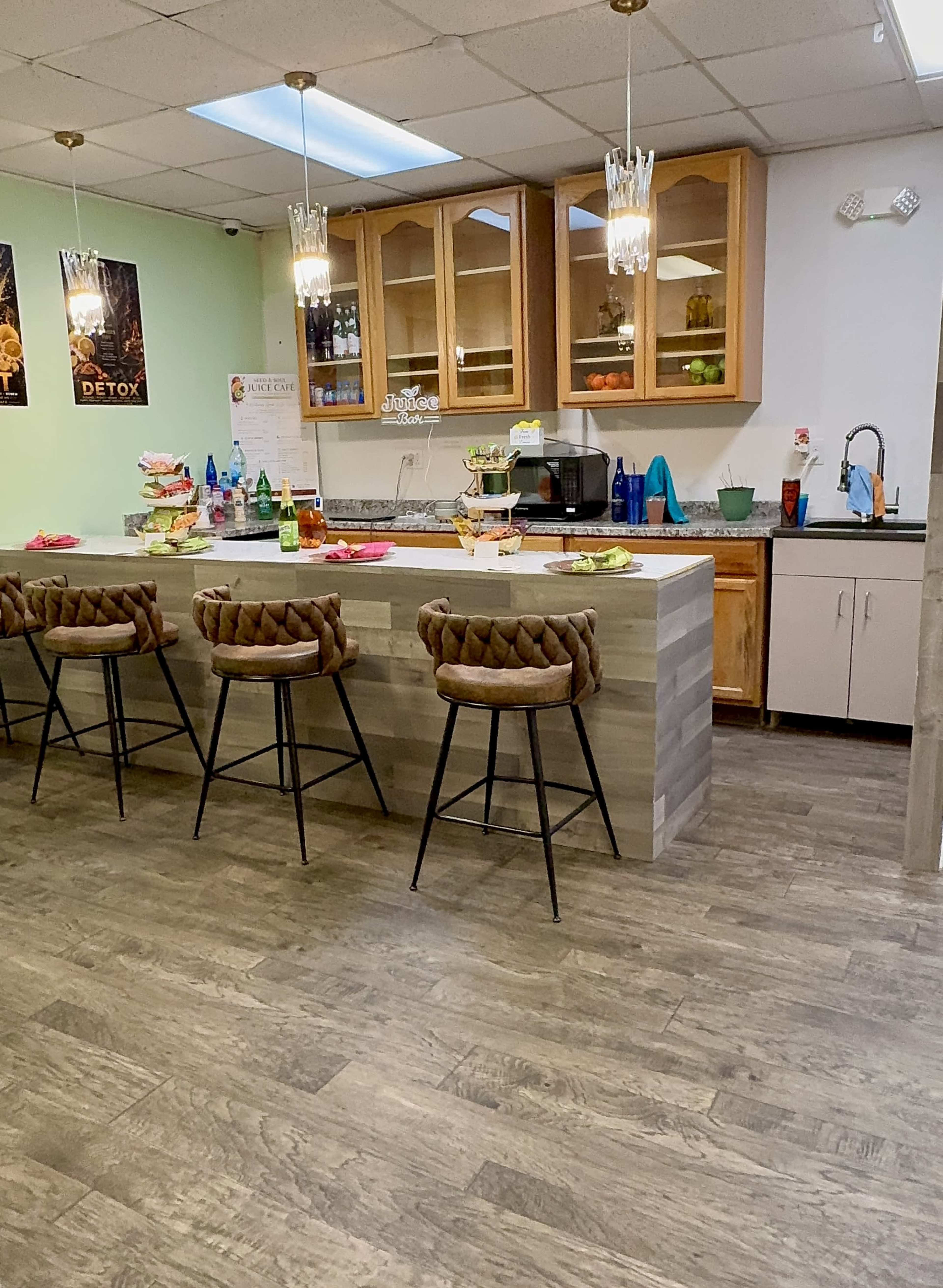 The image shows a modern kitchen bar area with high stools, a countertop filled with various drinks and snacks, and cabinets displaying colorful glassware.