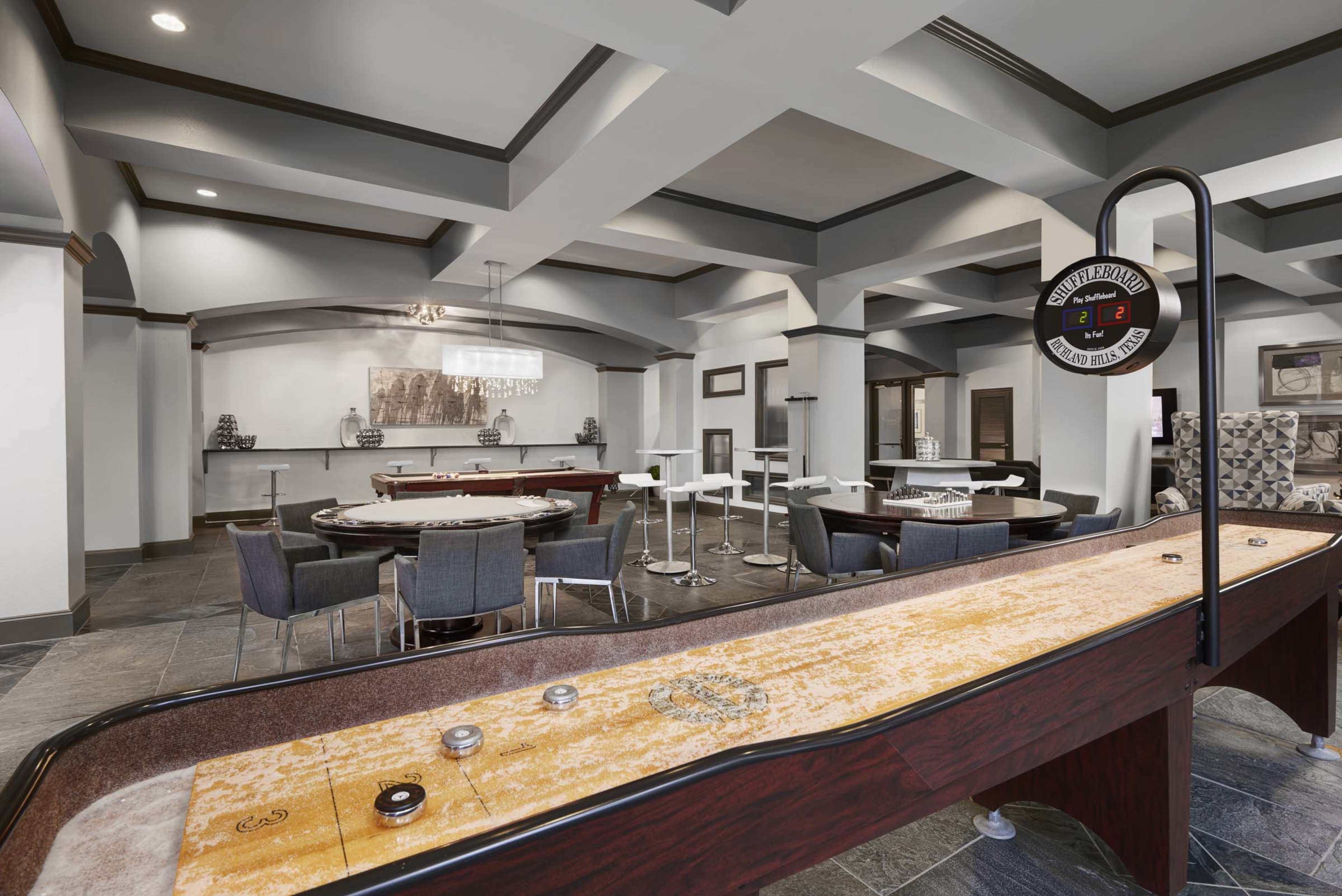 A spacious game room featuring a shuffleboard table, several card tables, and seating arrangements in a modern, well-lit setting.