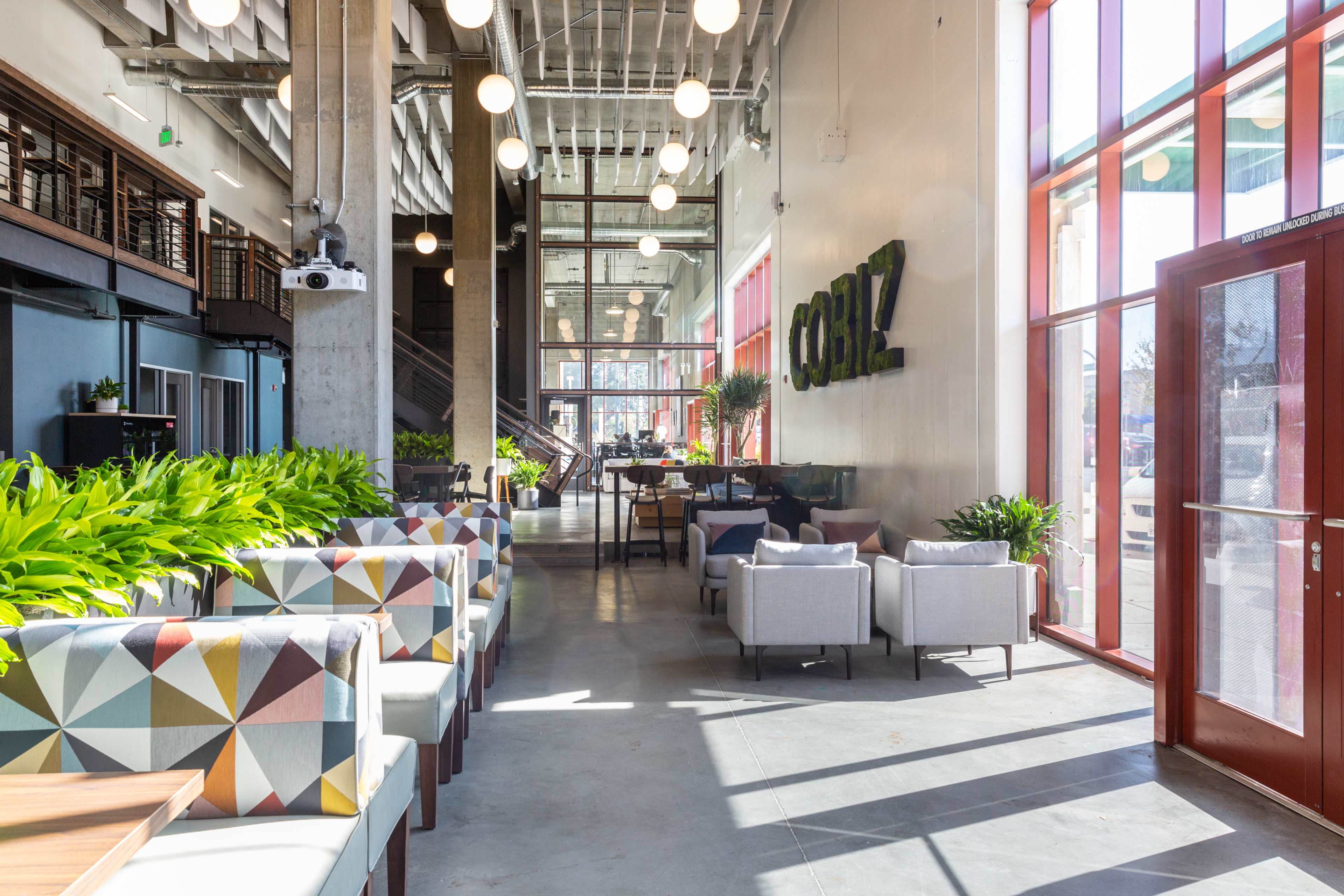 A modern café interior with colorful seating, large windows, and greenery throughout.