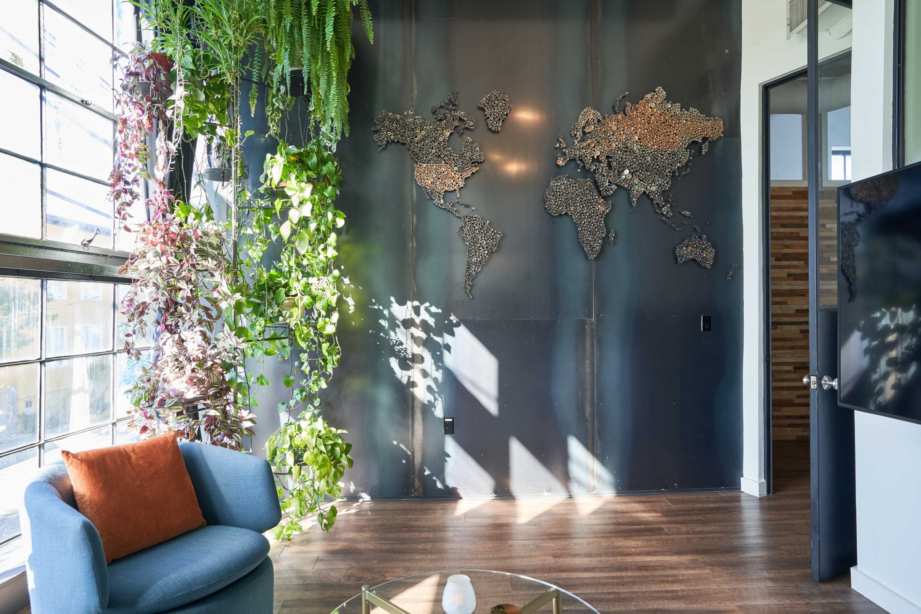 A modern indoor space featuring a world map made of metal on a dark wall, surrounded by hanging plants and a blue armchair with an orange cushion.
