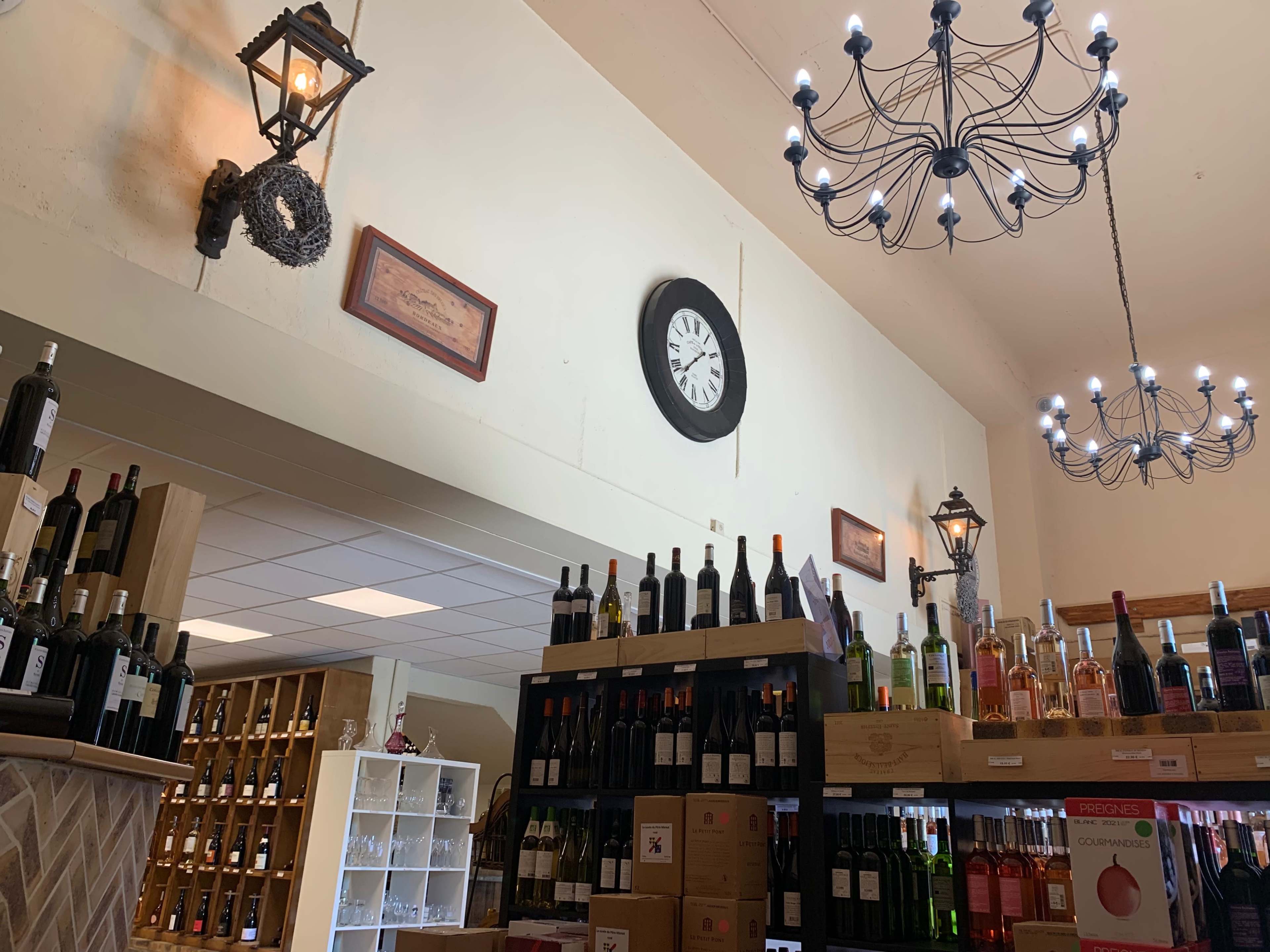 A wine shop interior with shelves of bottles, a clock on the wall, decorative lamps, and a chandelier overhead.