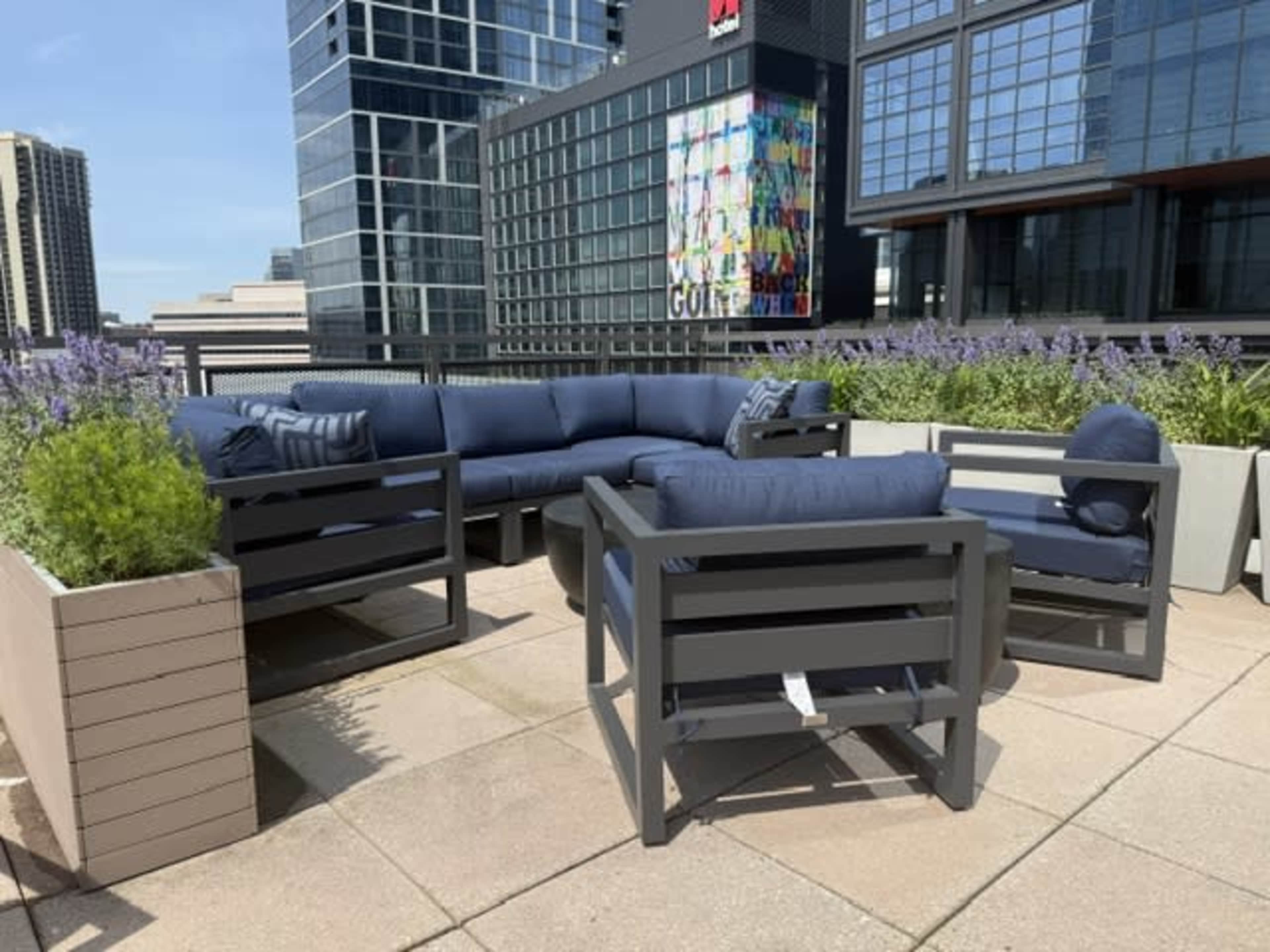 The image shows an outdoor seating area on a rooftop, featuring a sectional couch and chairs arranged around a patio space with planters filled with greenery.