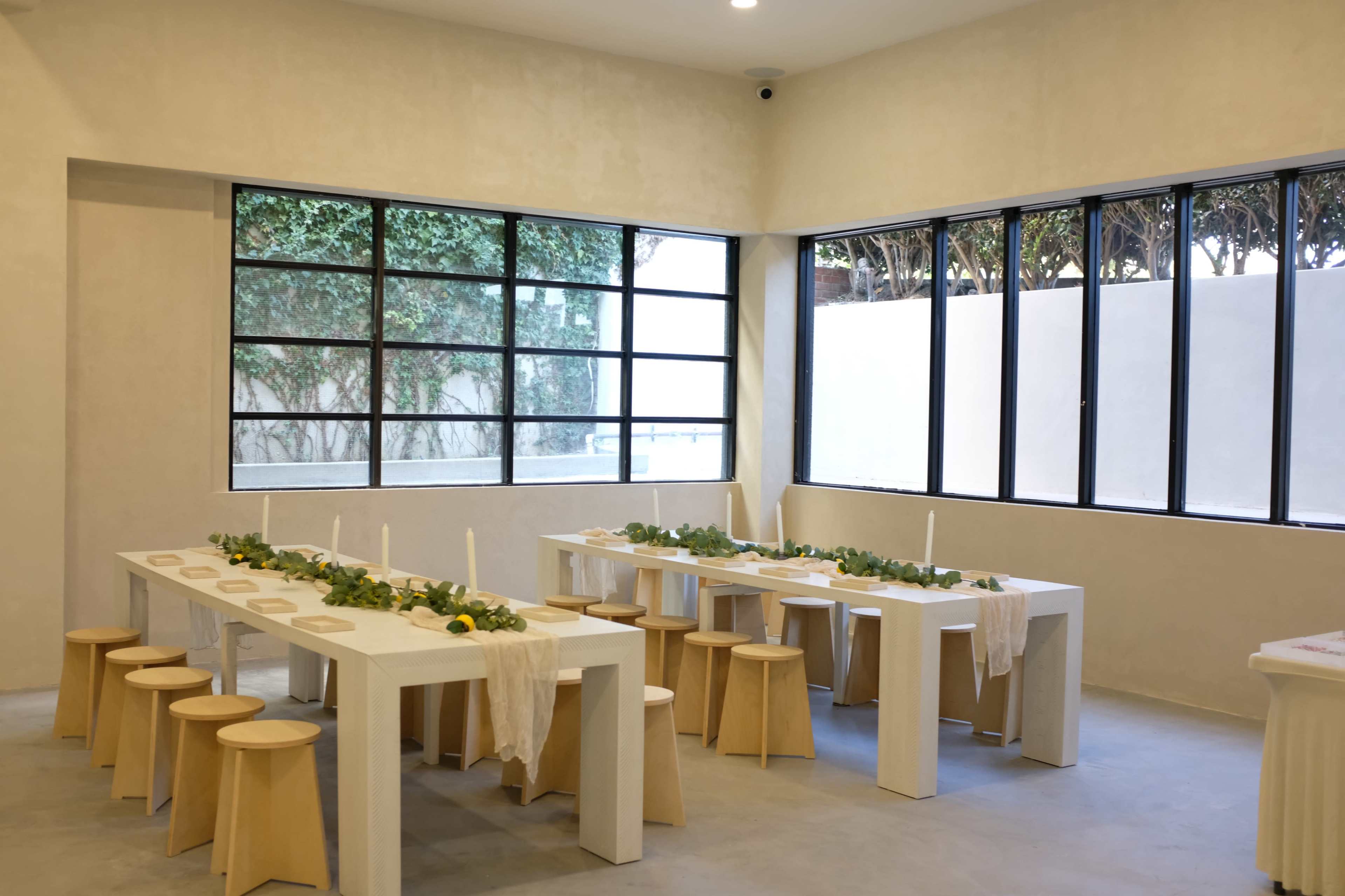 The image shows a minimalistic dining area with white tables arranged in two long lines, decorated with greenery and positioned near large windows that let in natural light.