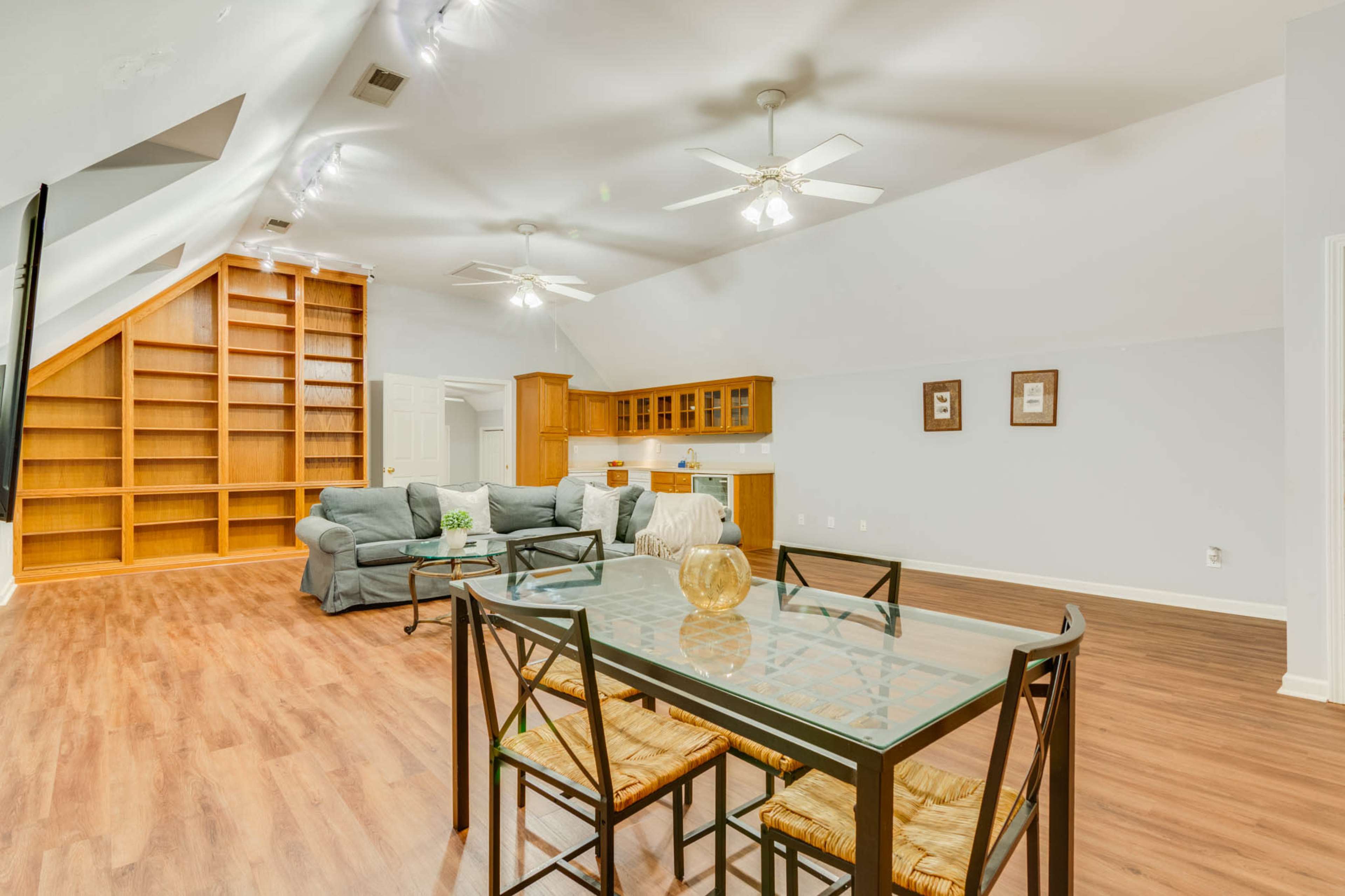The image shows a spacious room with a dining area featuring a glass table and chairs, and a living area with a couch, kitchen cabinetry, and a large bookshelf along one wall.