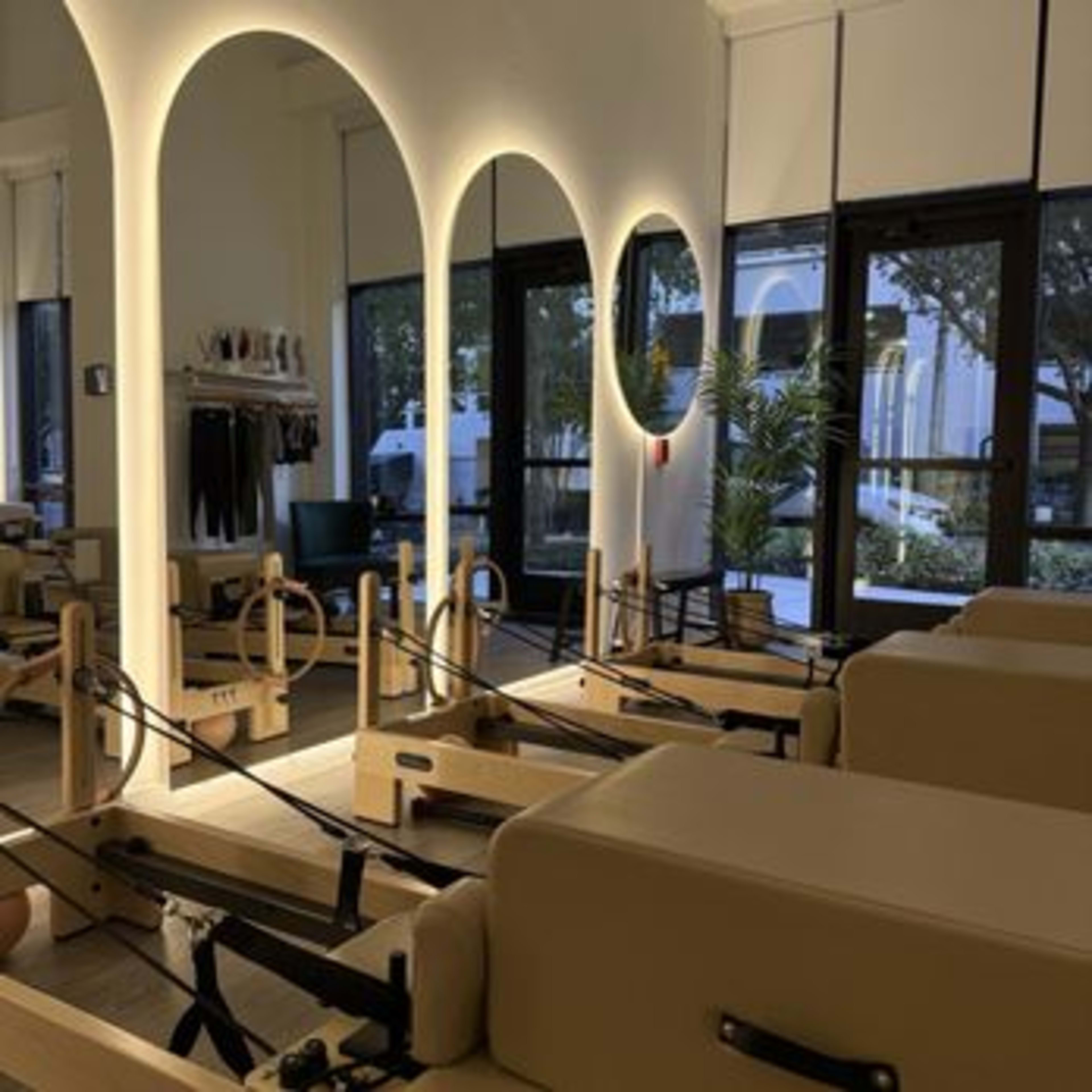 The image shows a bright and spacious studio with multiple Pilates reformer machines arranged under arched light fixtures.