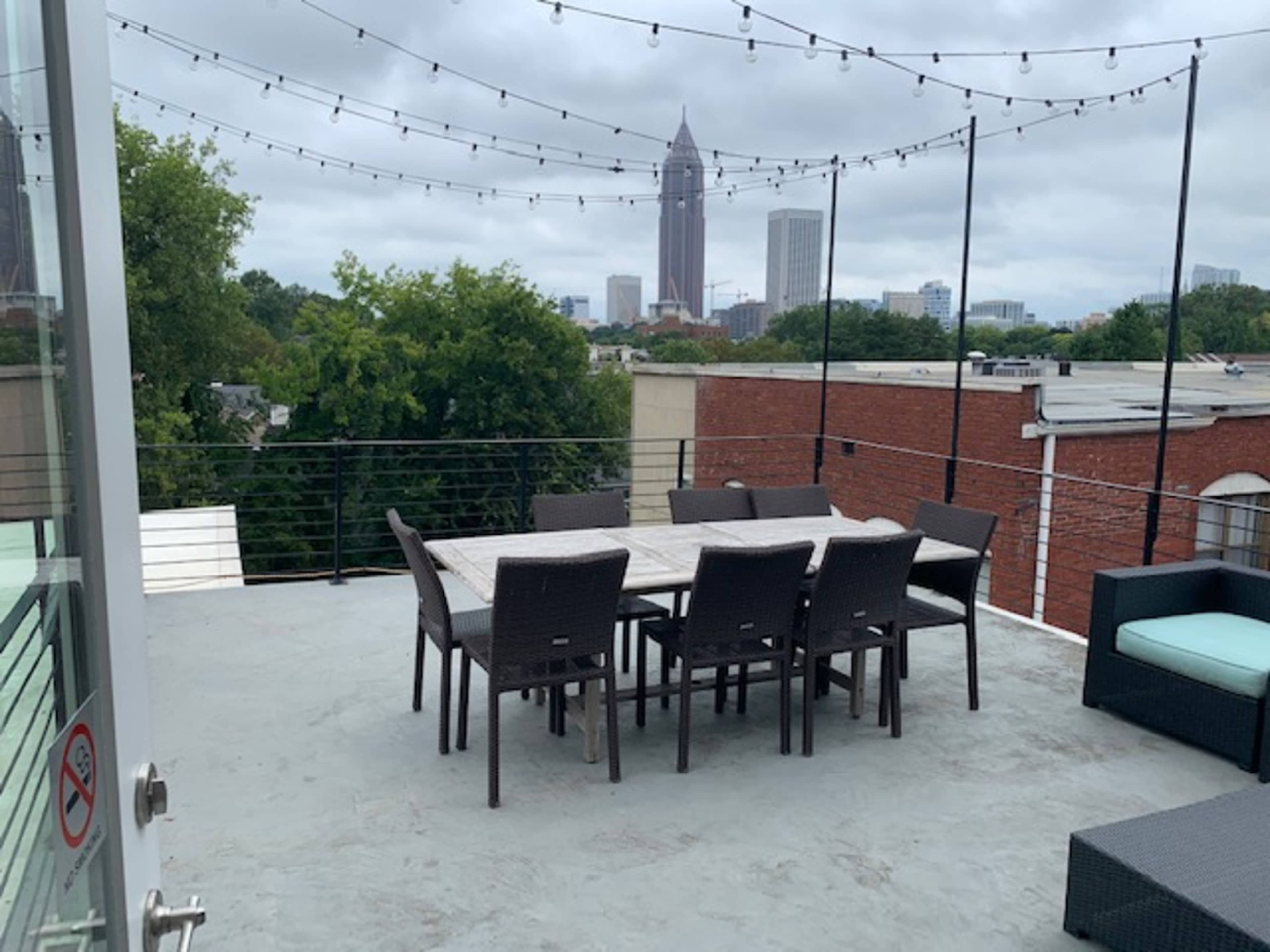A rooftop patio features a dining table surrounded by chairs and a view of tall buildings in the background under a cloudy sky.
