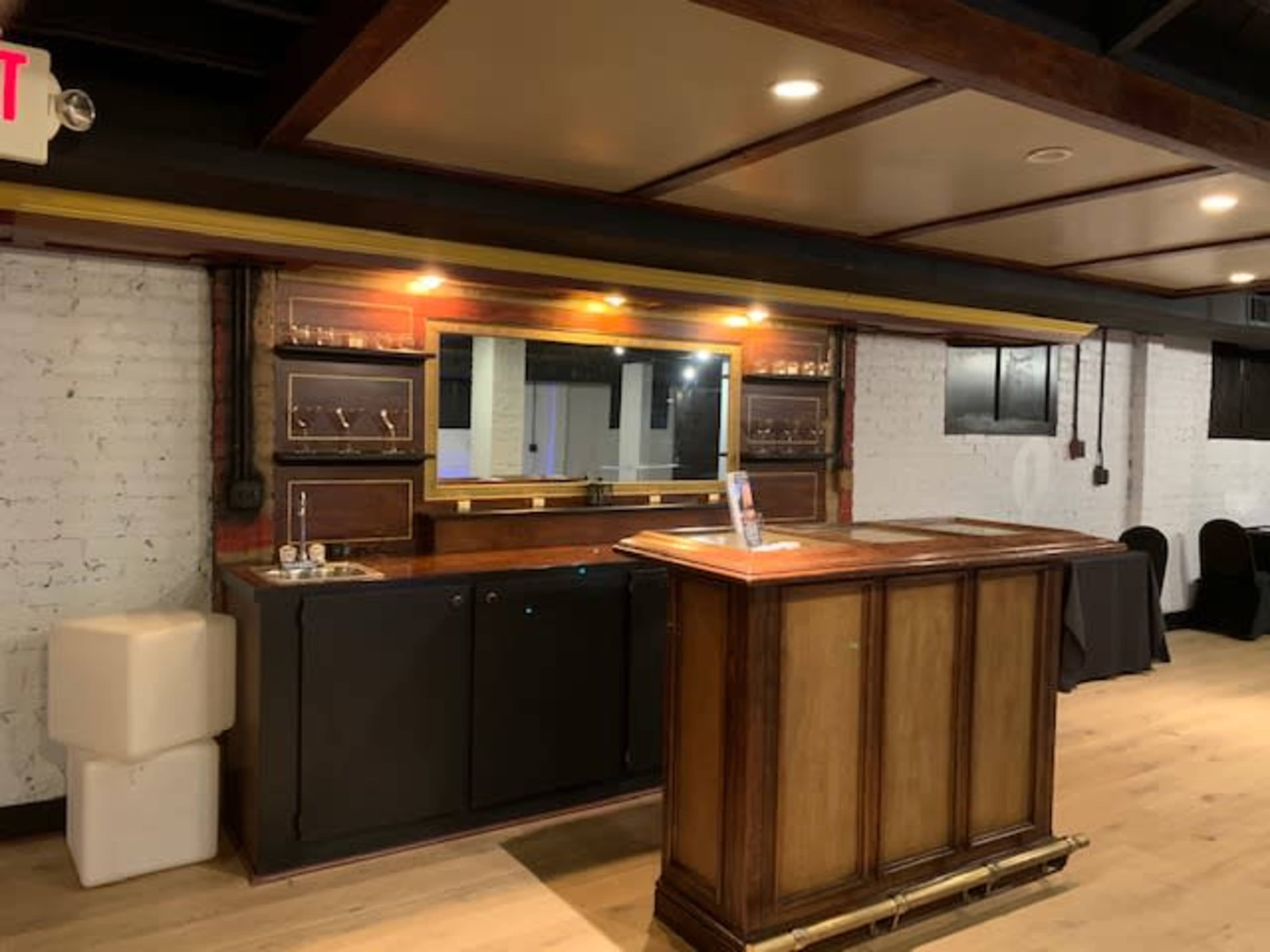A wooden bar with a mirrored back and shelves, set against a white-brick wall in a well-lit room.