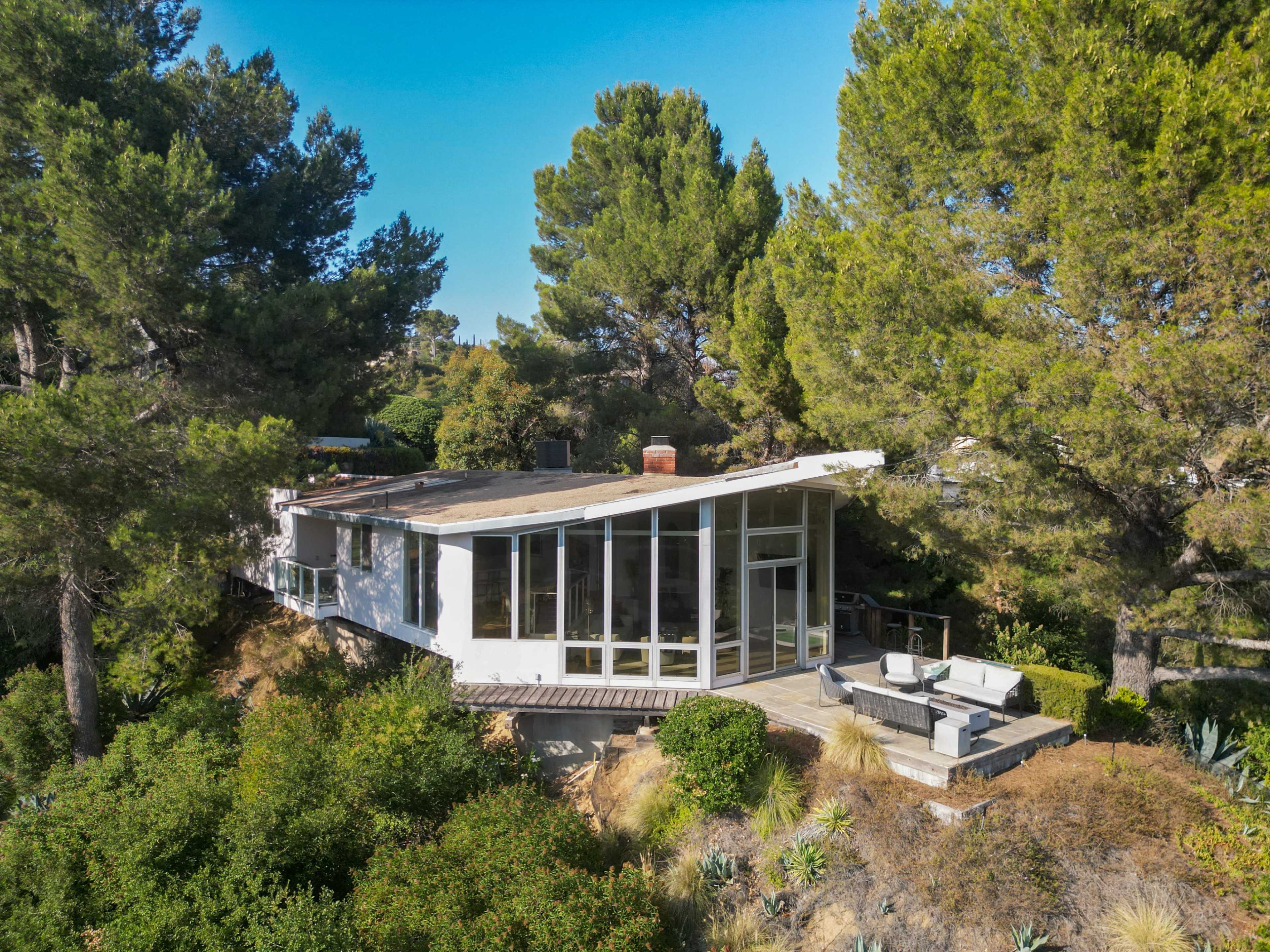 The image shows a modern house with large windows and a flat roof, situated among tall trees and greenery on a hillside.