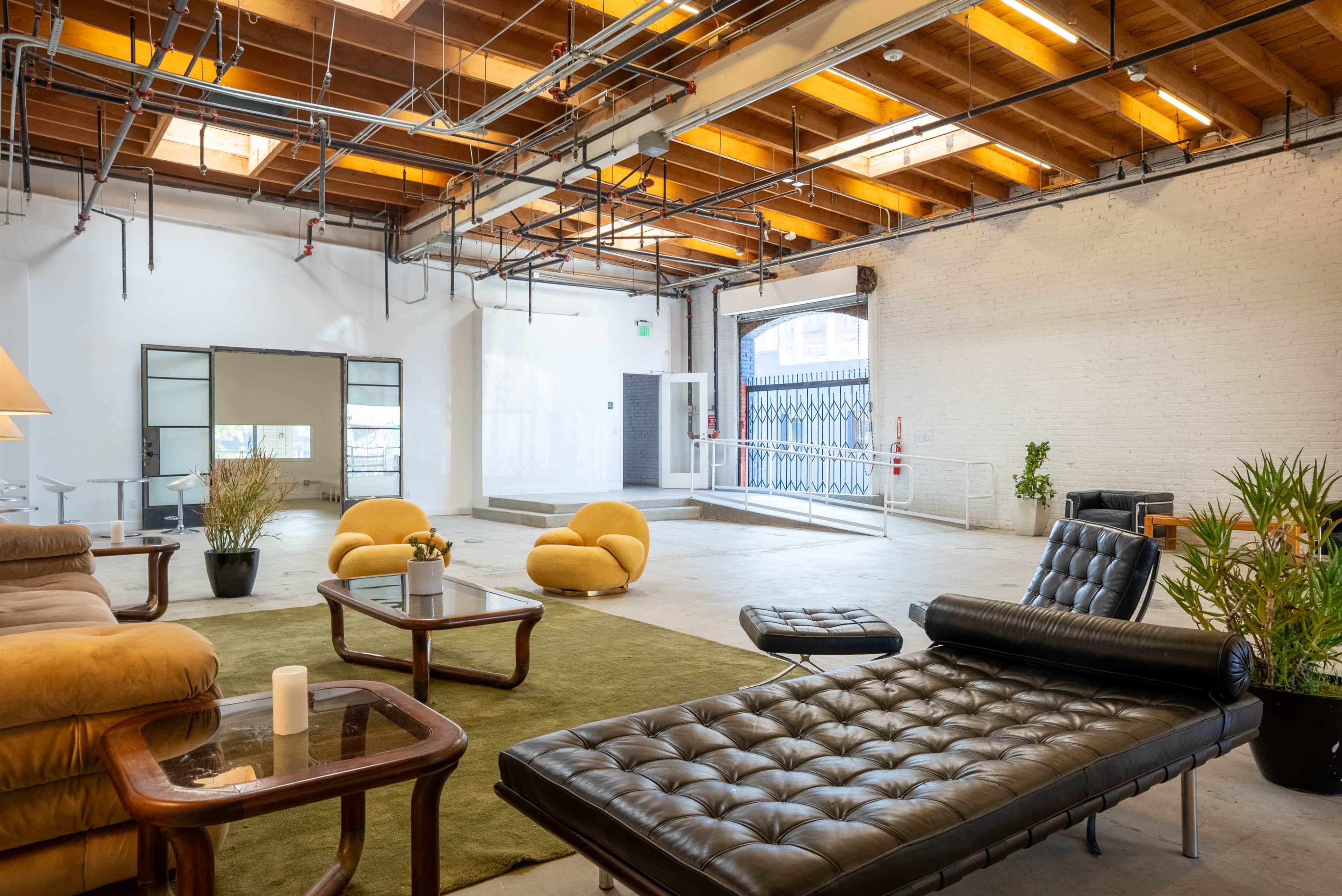 An open, spacious interior features modern furniture, including a black chaise lounge, a brown leather sofa, and round yellow poufs on a green carpet.