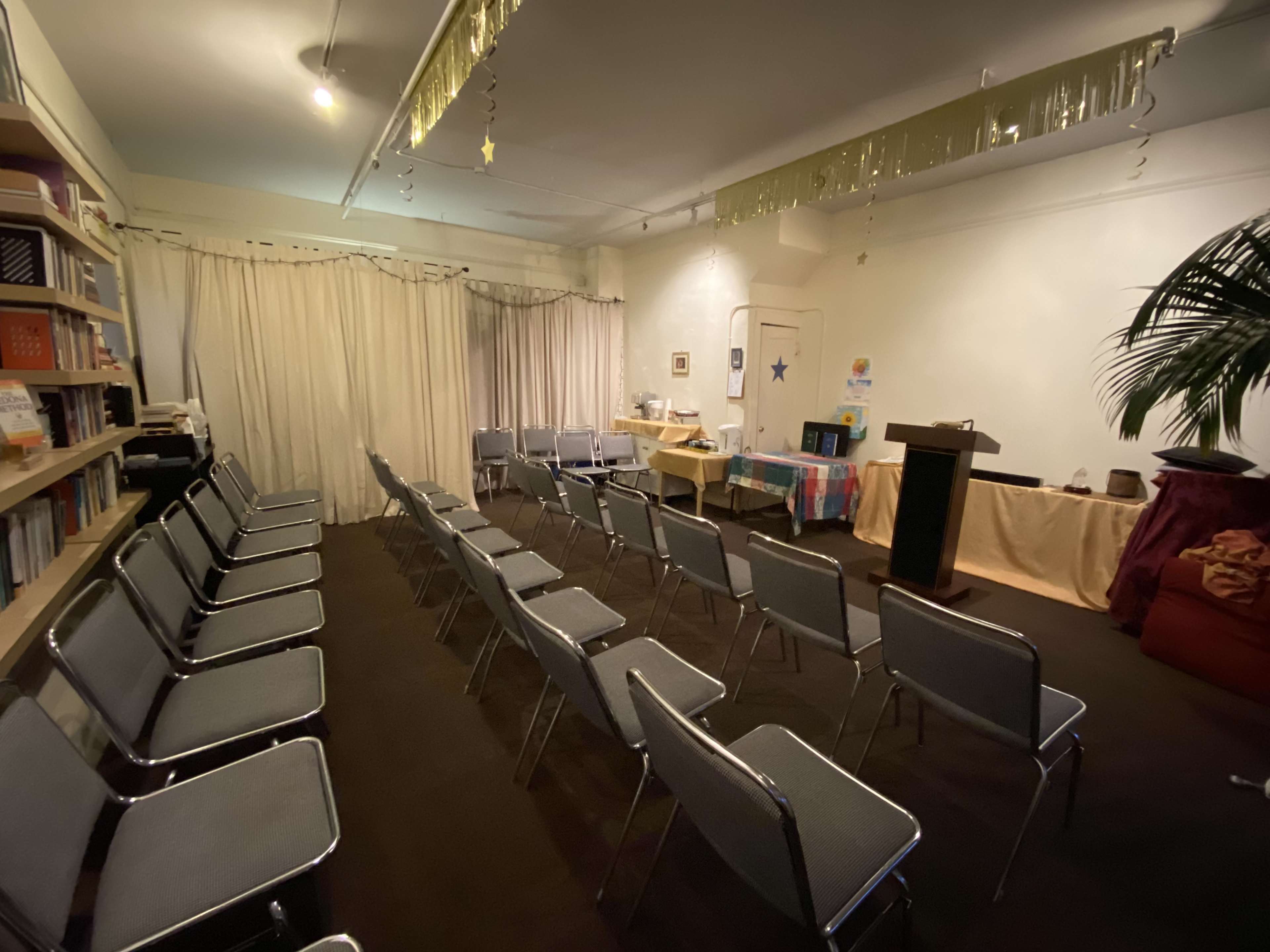 A room with neatly arranged metal chairs facing a small podium, surrounded by tables and decorations, including a large curtain and a potted plant.