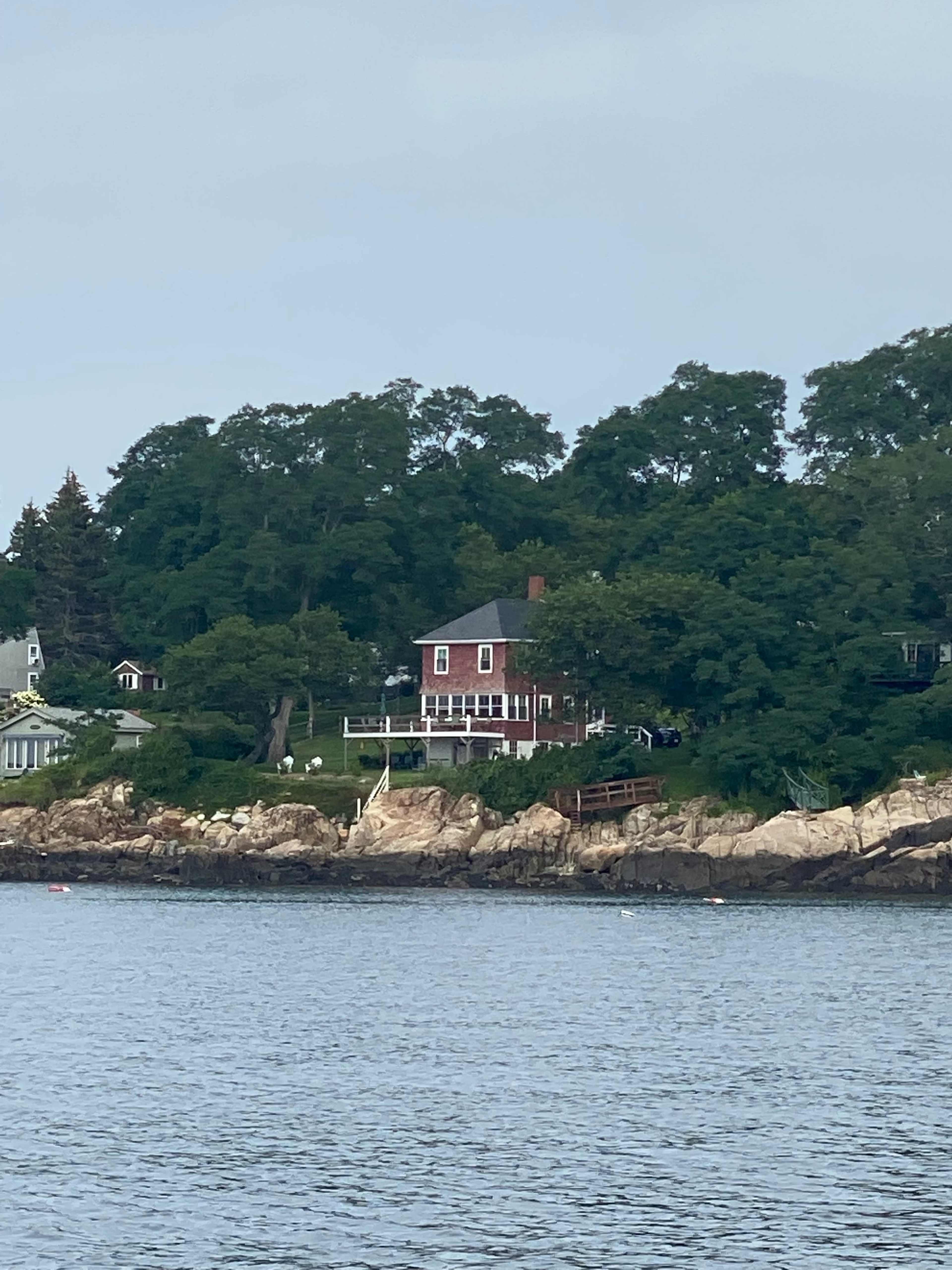 A red house is situated on a rocky shoreline, surrounded by trees and overlooking the water.