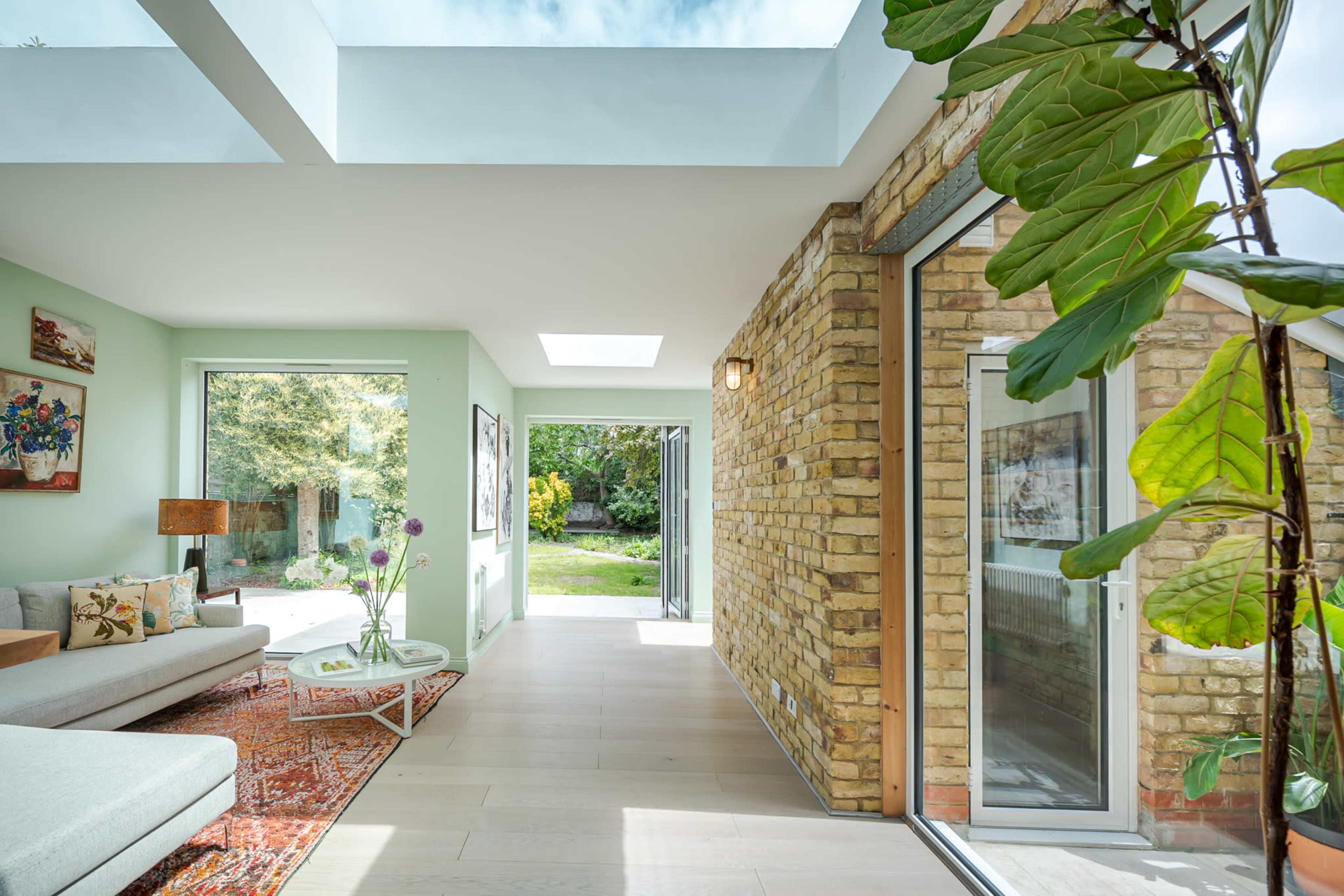 The image shows a modern living room with large windows, a brick wall, and a view of a garden.