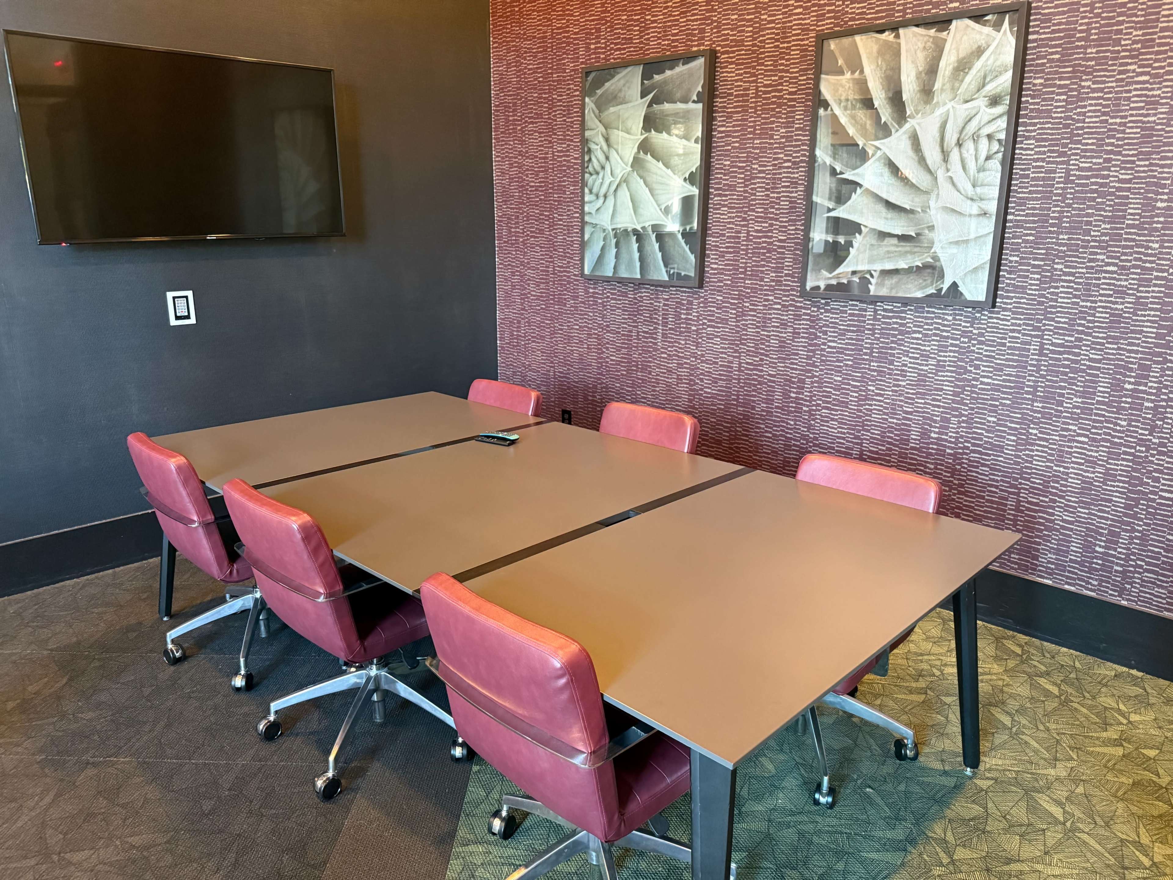 A conference room with a large table surrounded by six red chairs, featuring a television on the wall and two framed artworks.